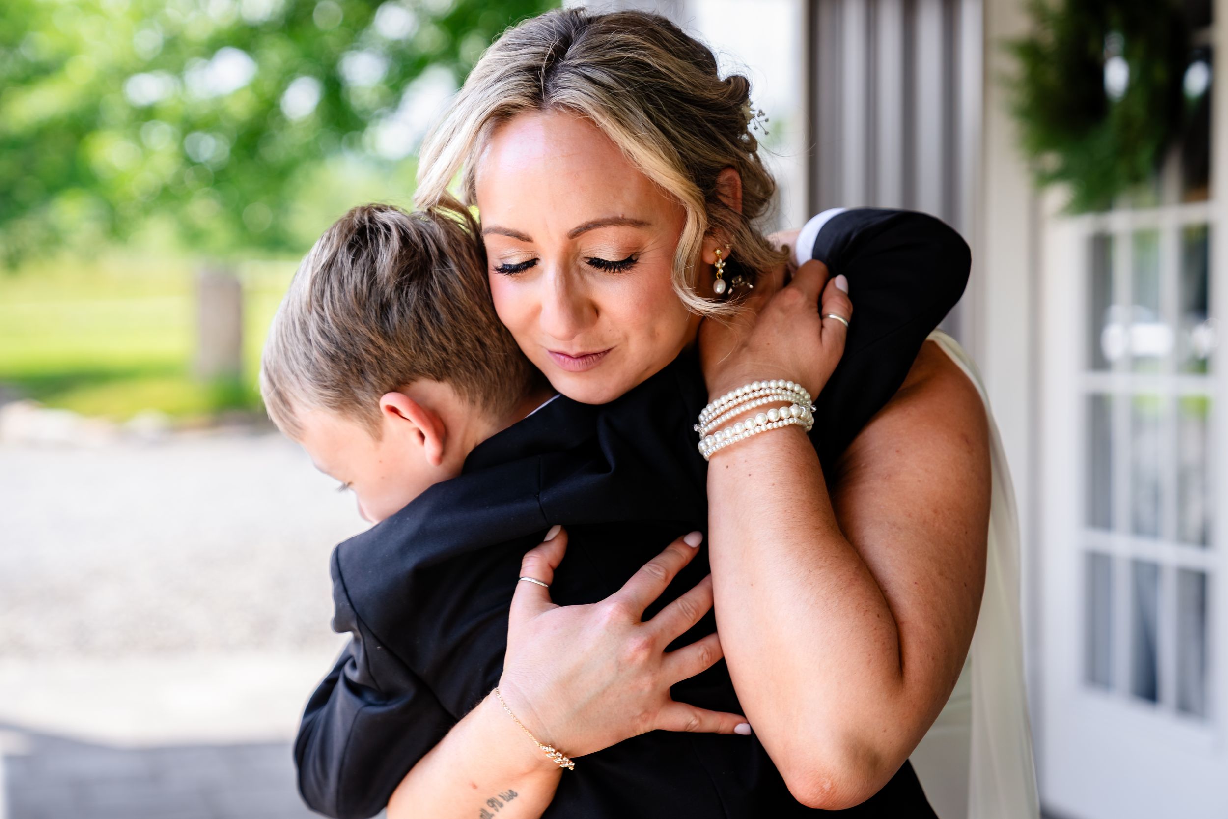 Bride in black dress sharing a tender hug with a young boy in a suit at an outdoor wedding venue.