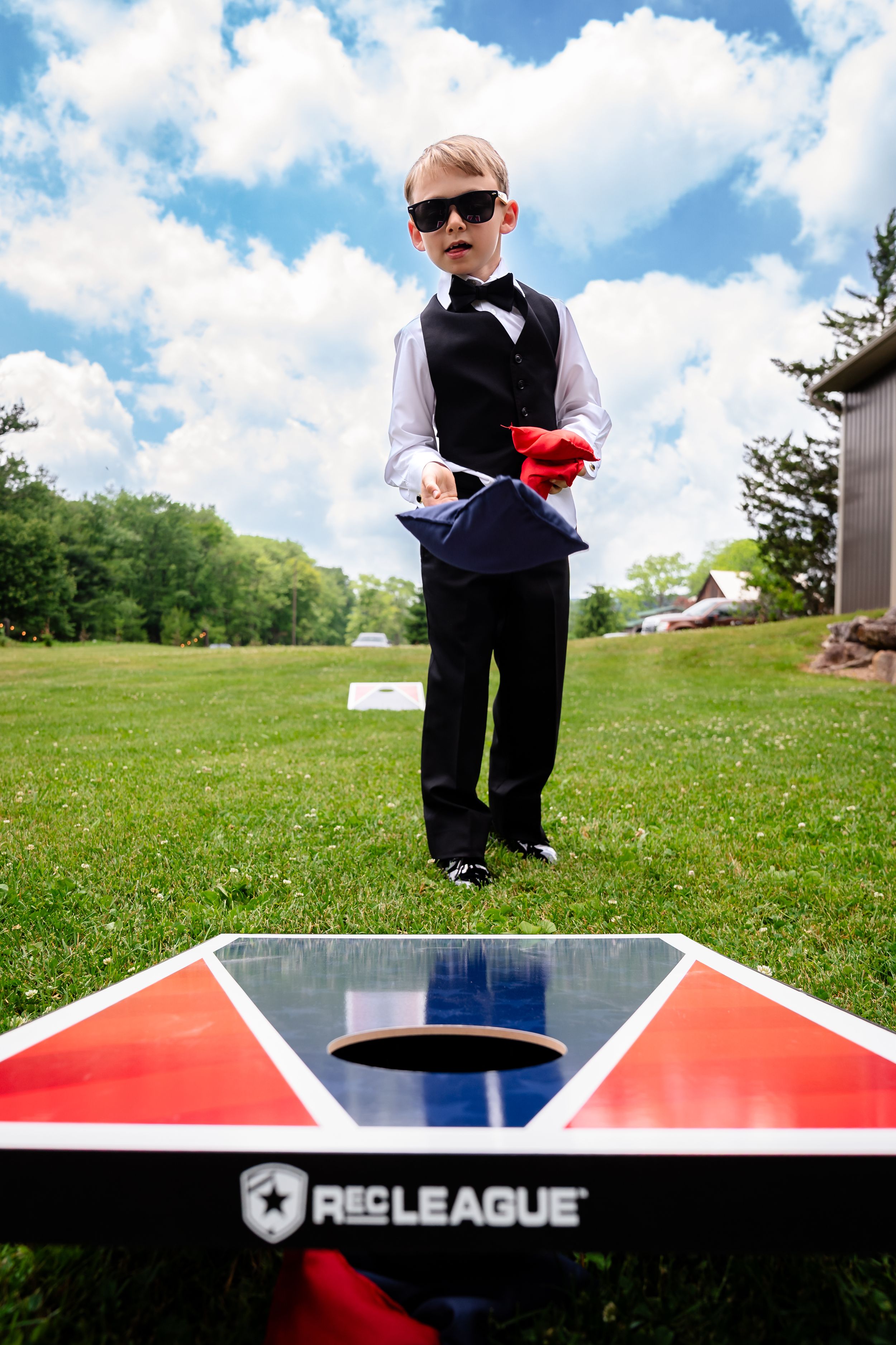 Stylish young ring bearer in sunglasses plays cornhole on sunny wedding lawn.
