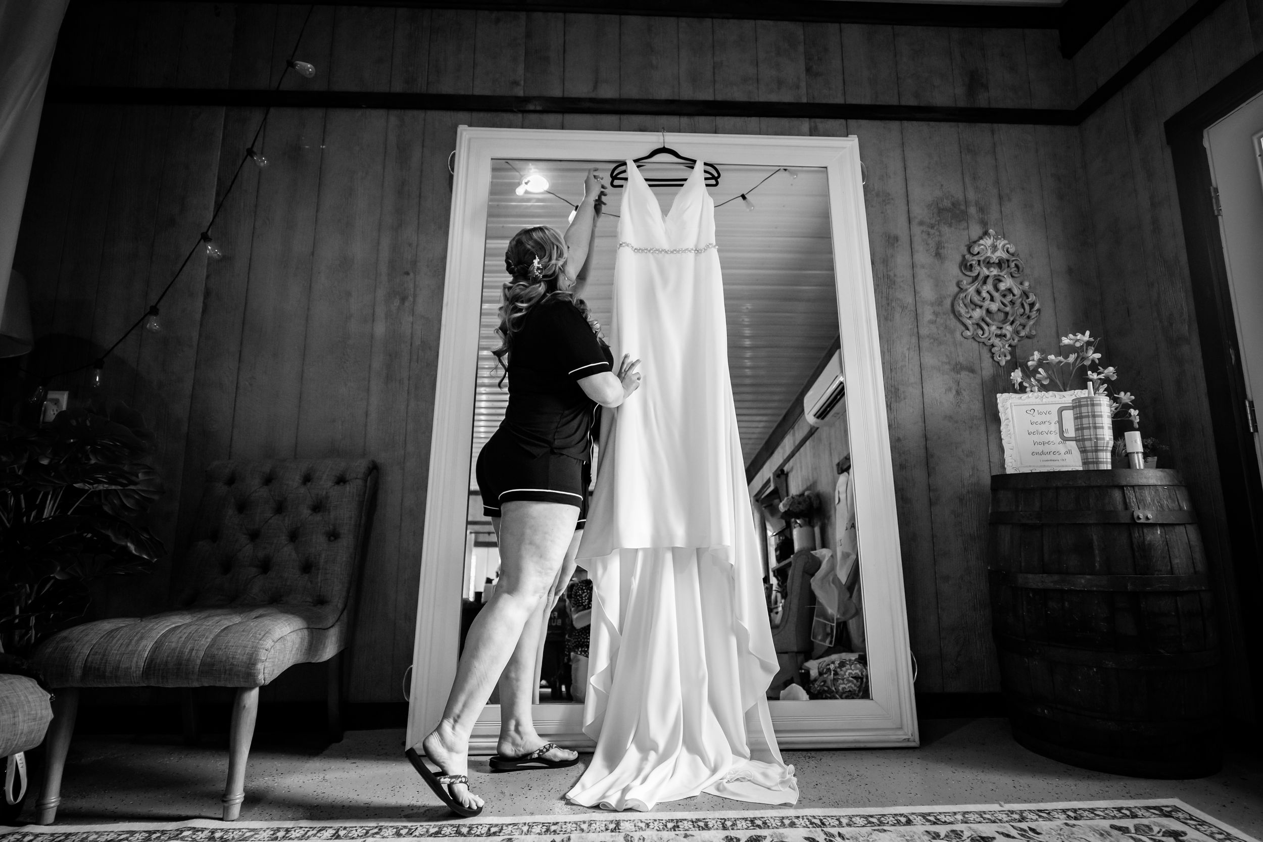 Black and white photo of a woman admiring a wedding dress hanging in a doorway of a rustic room.