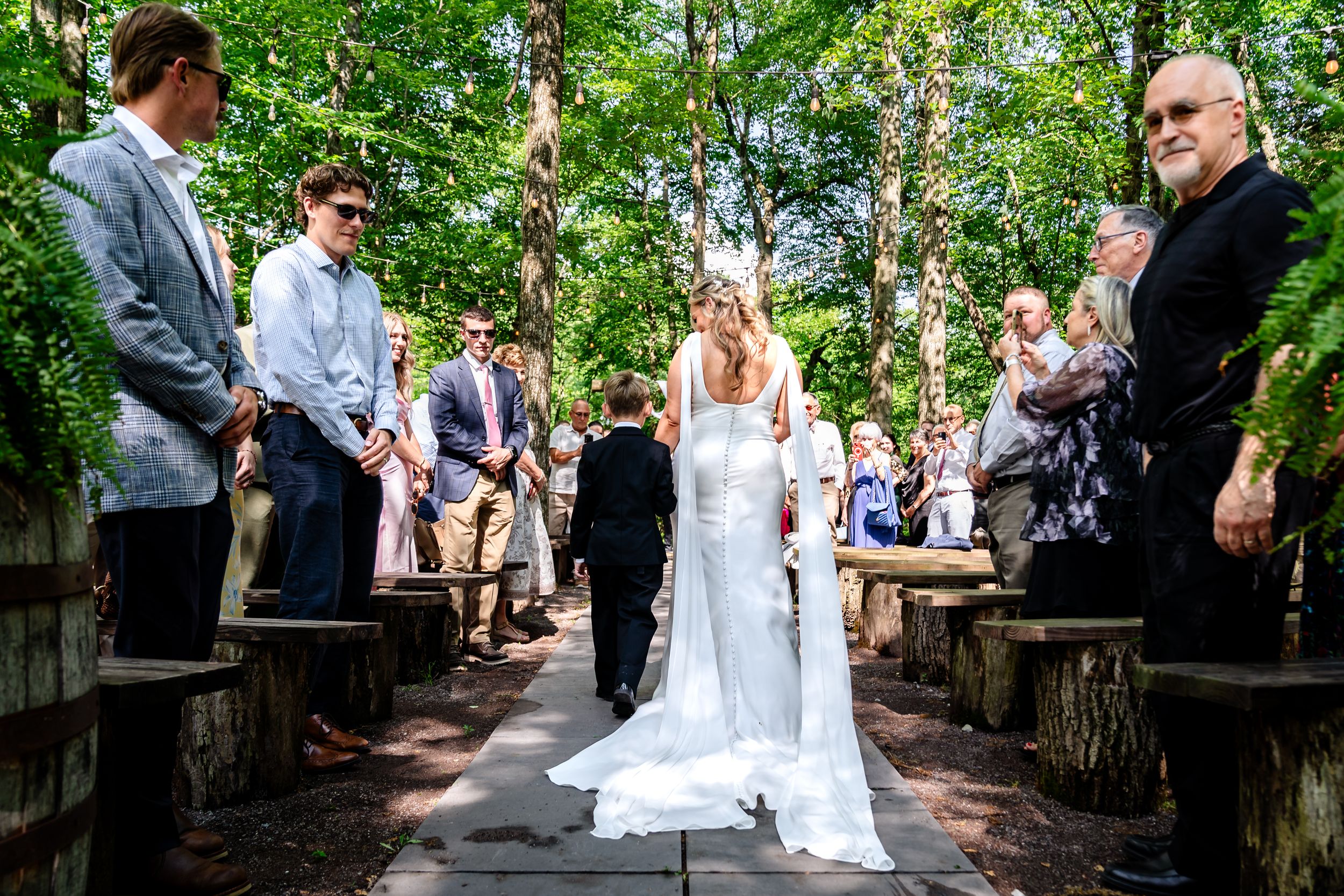 Bride walking down a wooded outdoor aisle in a white gown surrounded by wedding guests on a sunny day.