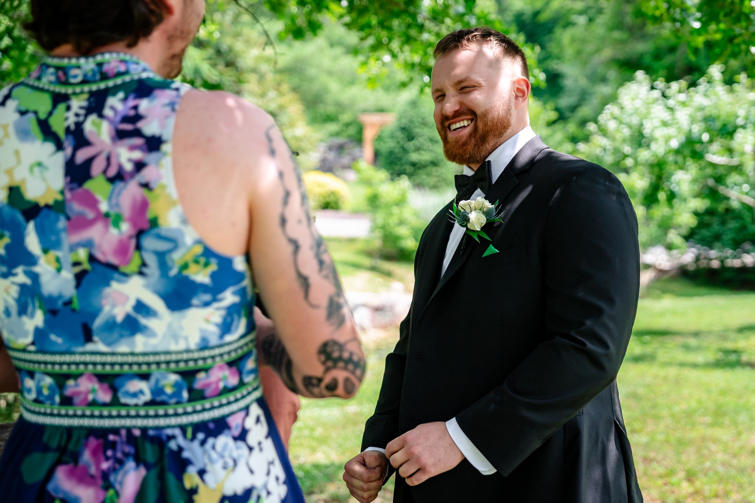 Smiling red-haired groom in black tuxedo with boutonniere during outdoor wedding ceremony.