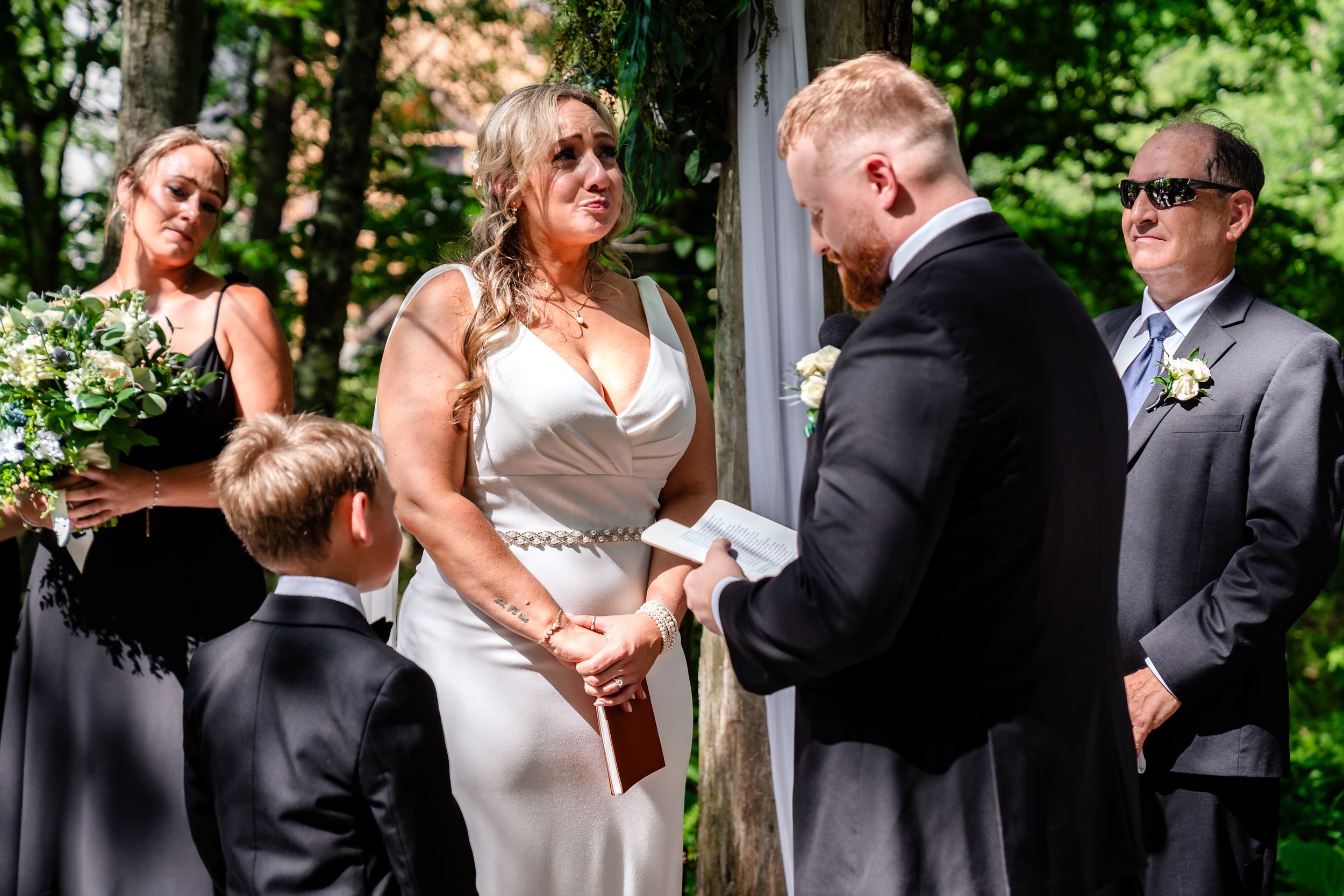 Bride in white gown exchanges vows with groom outdoors at elegant woodland wedding ceremony.