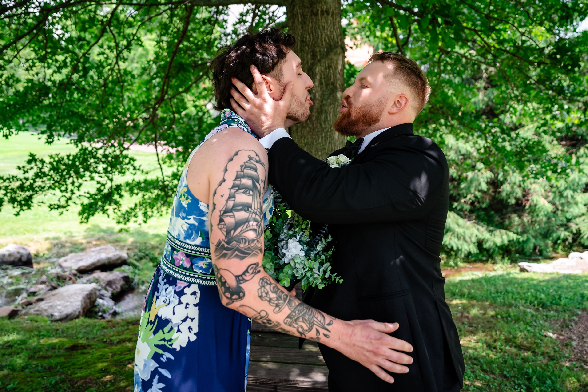 Tattooed couple sharing a kiss under a lush green tree during their outdoor wedding ceremony.