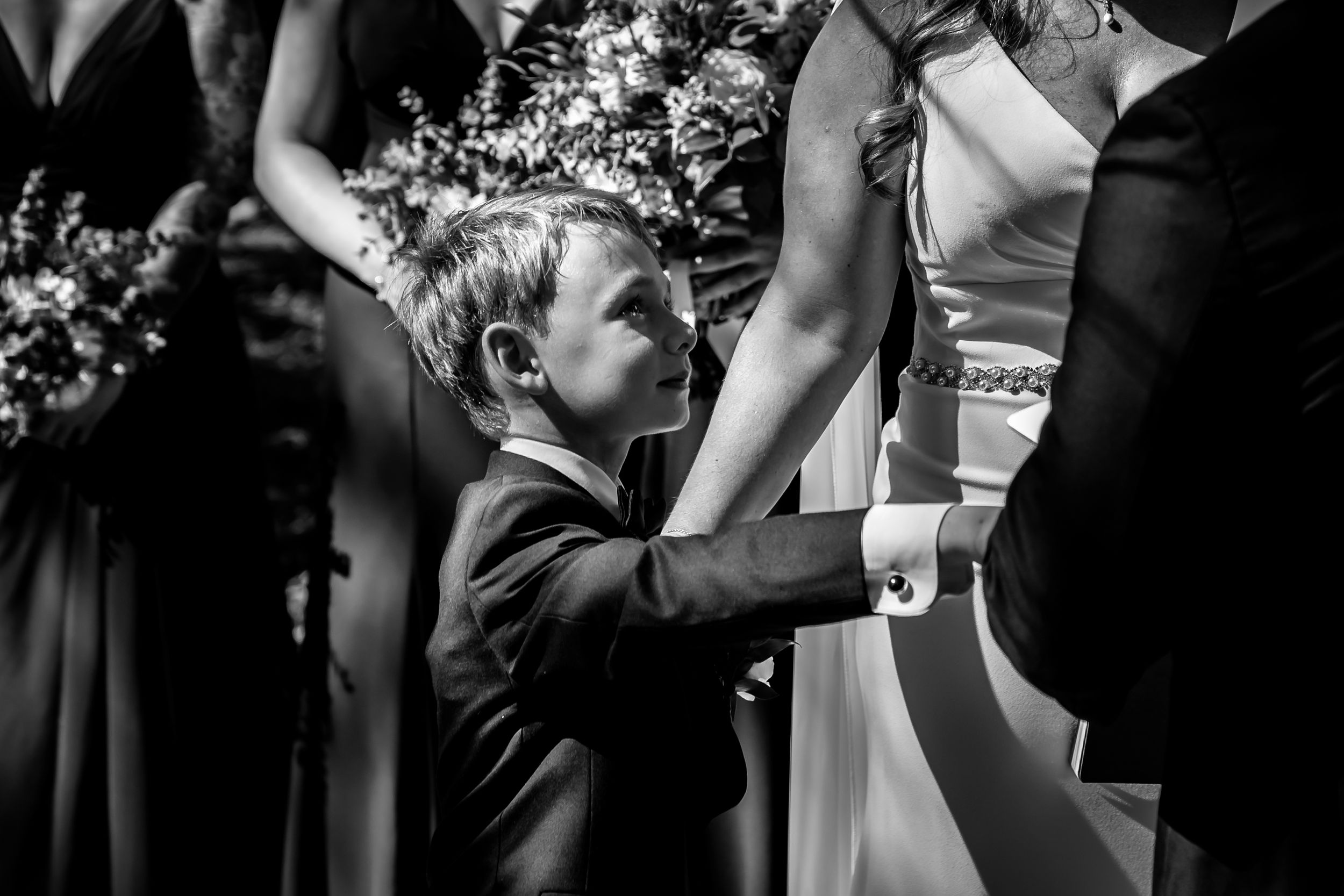 Young ring bearer in suit holds bride's hand during black and white wedding ceremony photo.