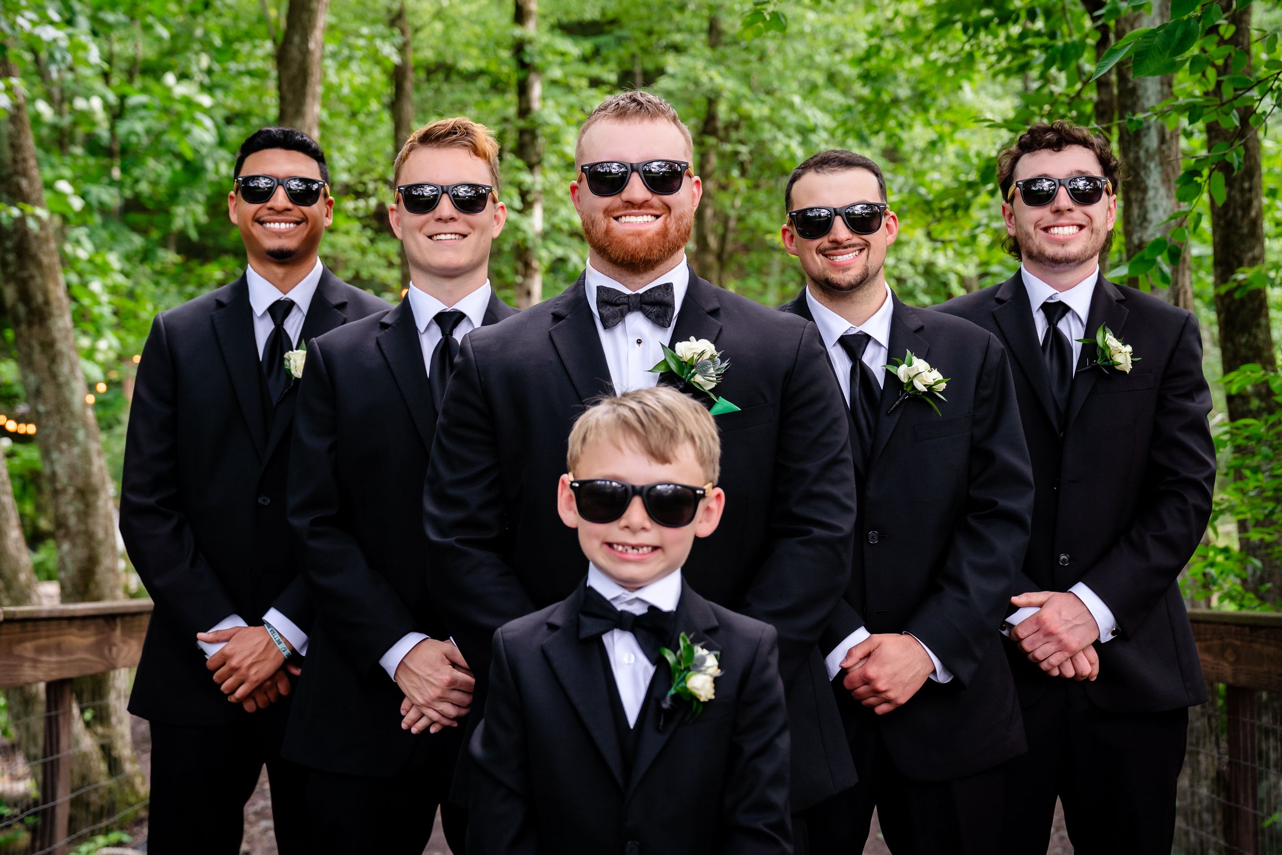 Groomsmen and ring bearer in black tuxedos and sunglasses pose outdoors in a wooded setting for a wedding photo.