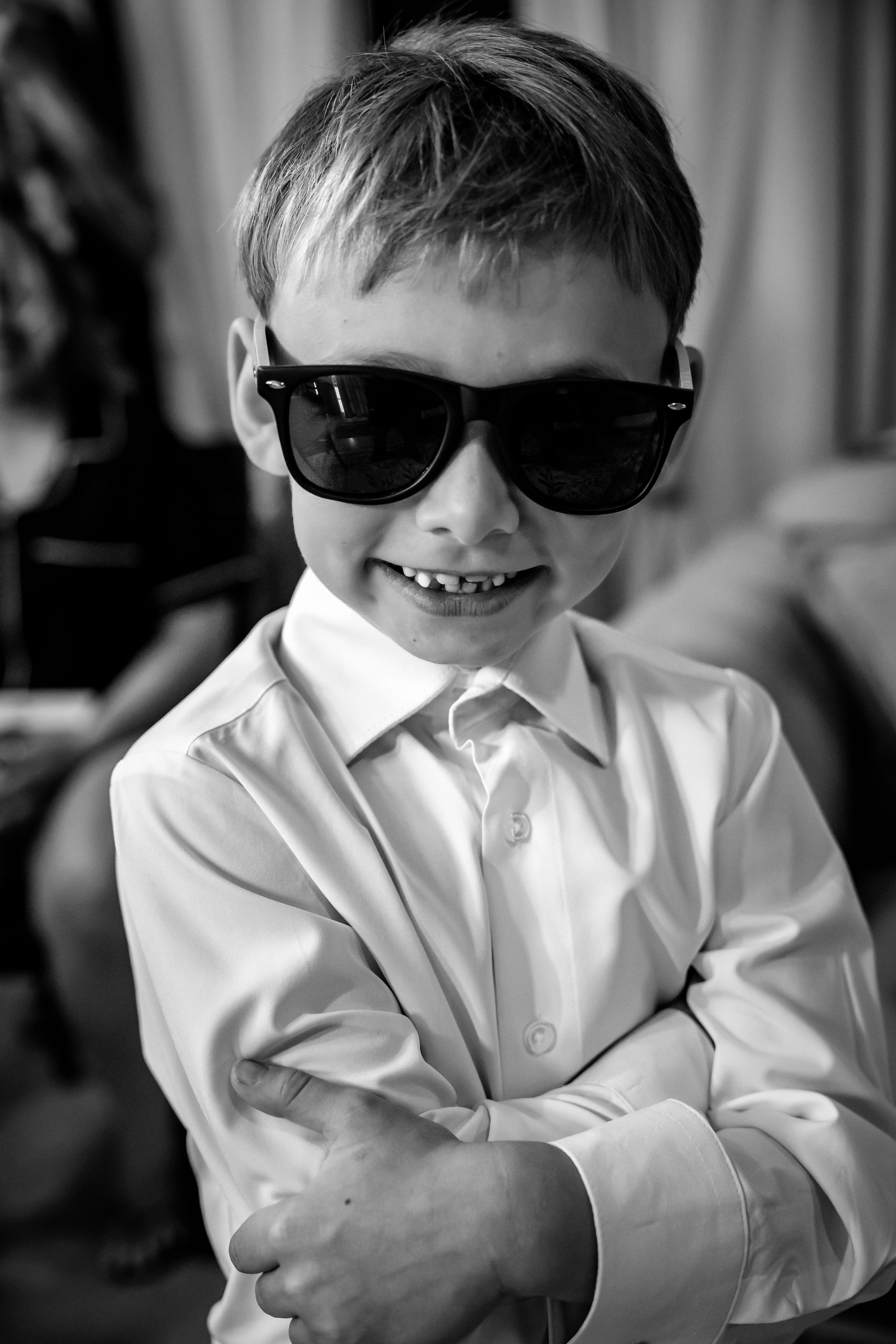 Adorable young boy in white shirt poses confidently wearing oversized black sunglasses in black and white photo.