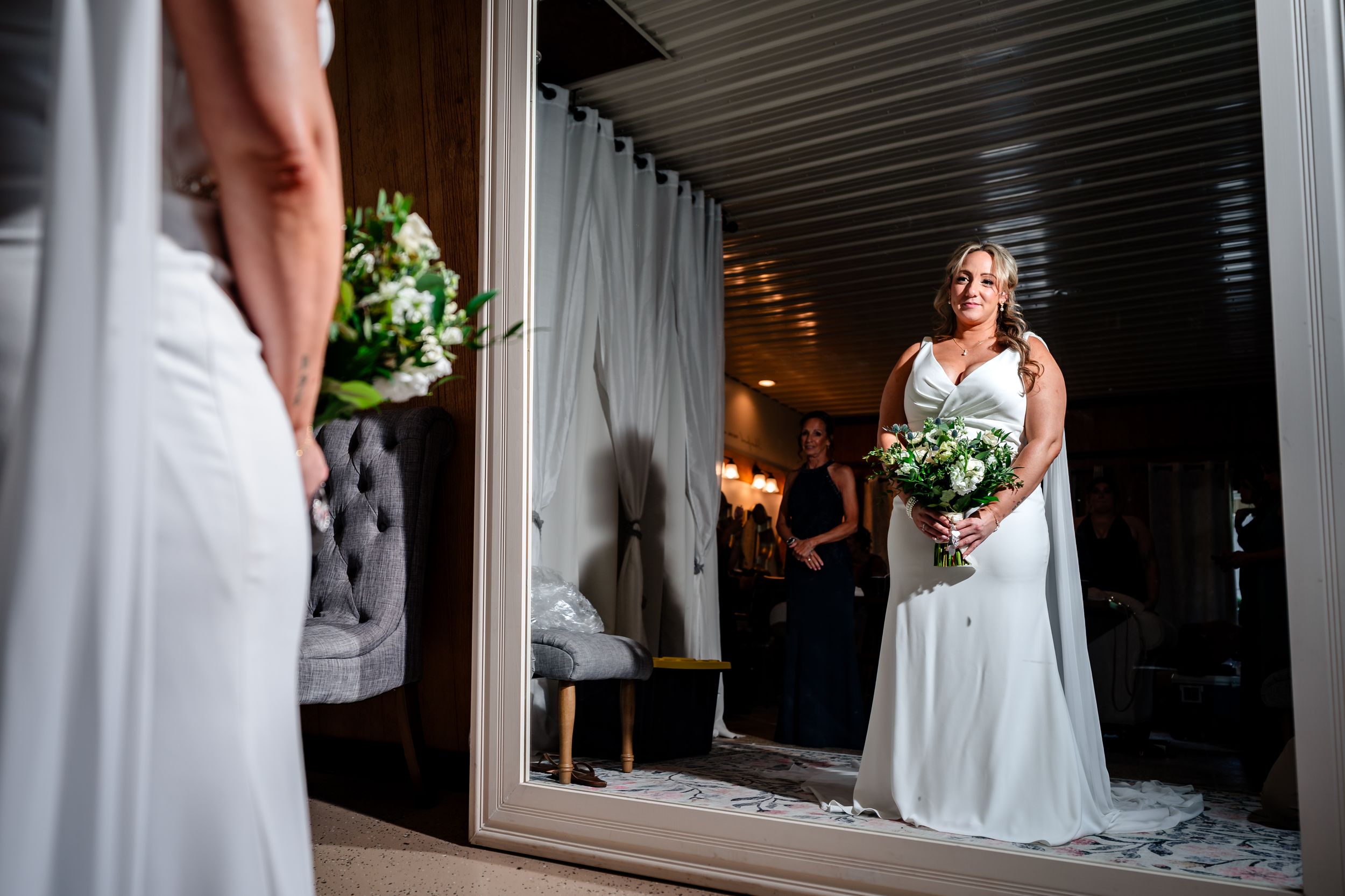 Bride in white gown holding bouquet reflected in mirror before wedding ceremony.