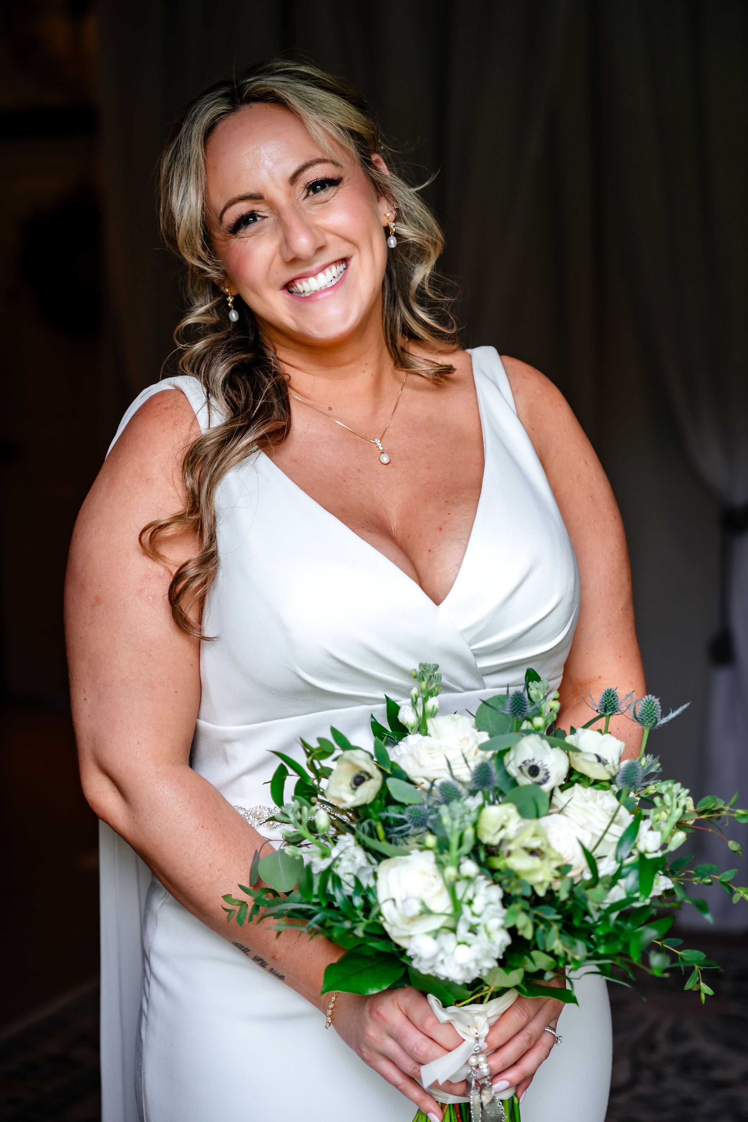 Smiling bride in white V-neck gown holding a bouquet of white flowers and greenery.