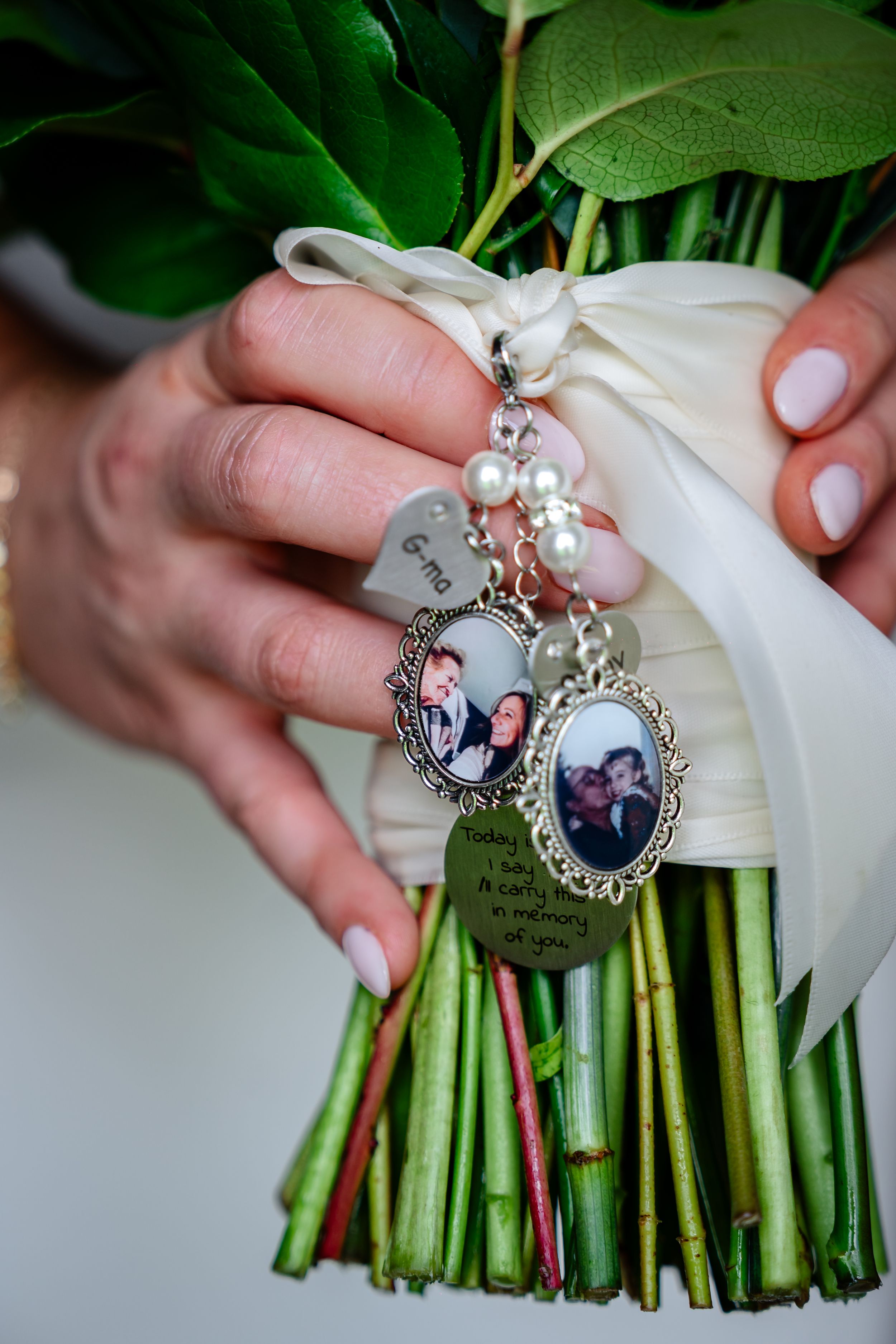 Bridal bouquet with memorial photo charms and pearl accents, held by bride with white manicure.