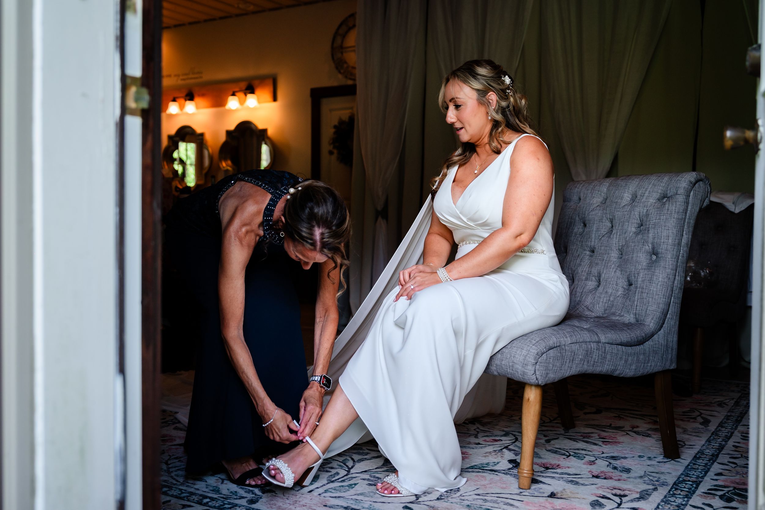 Bridesmaid kneels to fasten the brides pink heeled sandals as she sits on a gray tufted chair in a bridal suite.