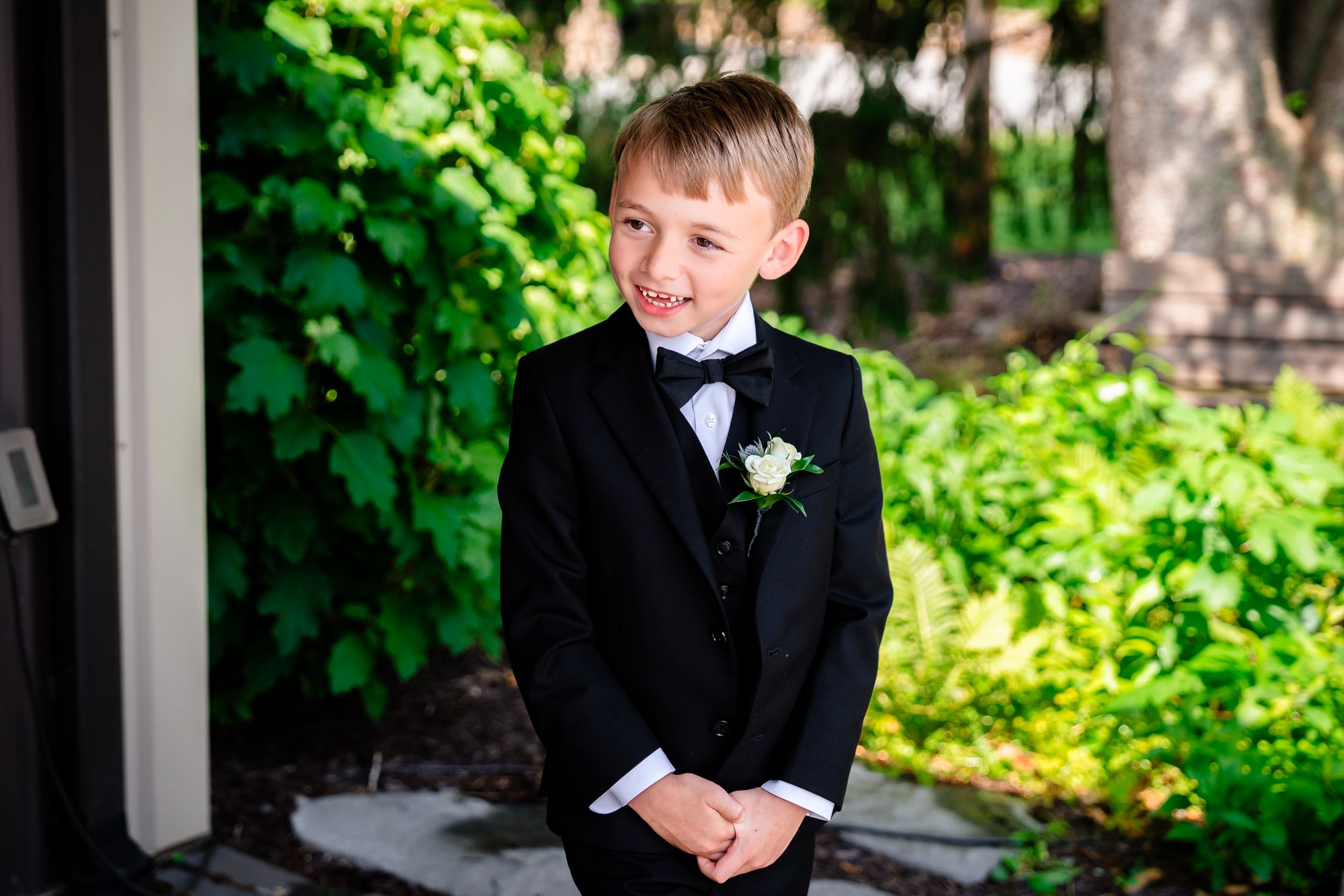 A young boy in a black tuxedo with a white boutonniere smiles outdoors.