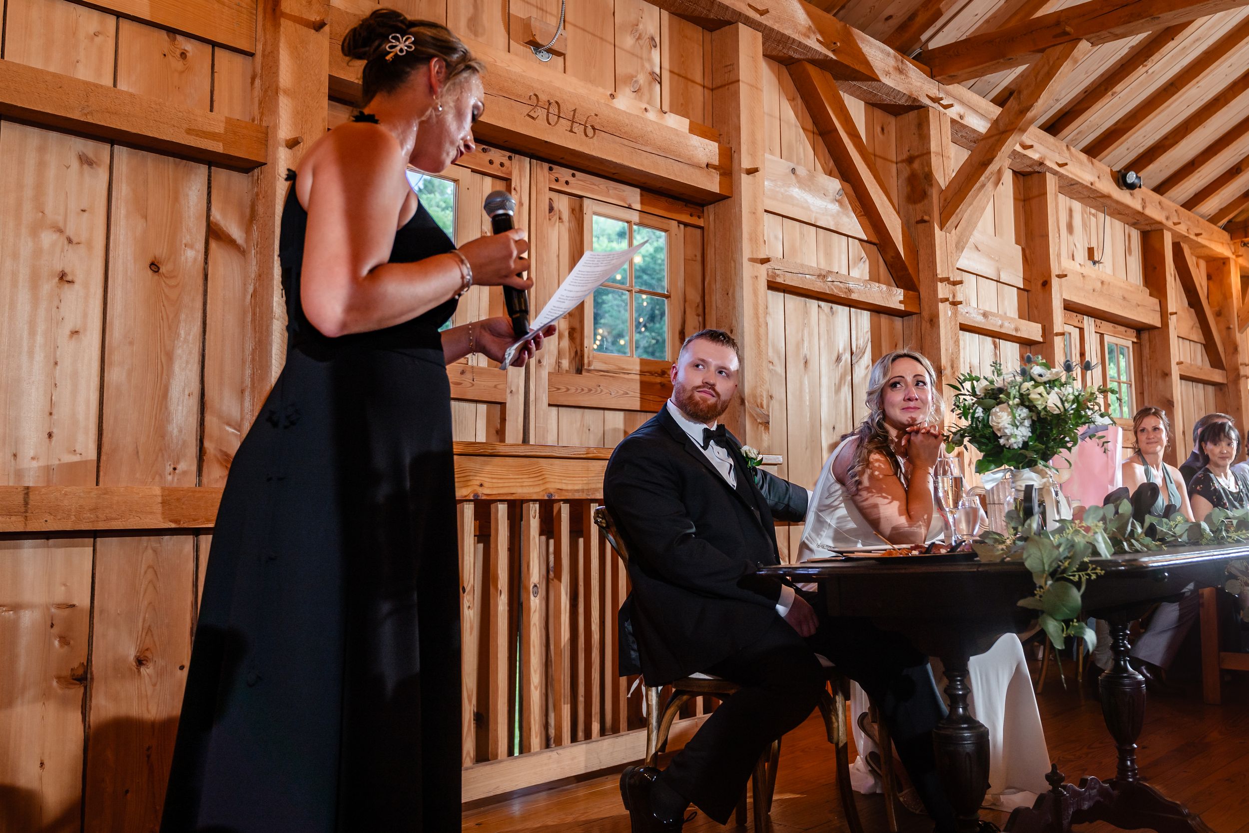 Woman in black dress giving wedding toast speech at rustic barn reception while guests listen at decorated table.