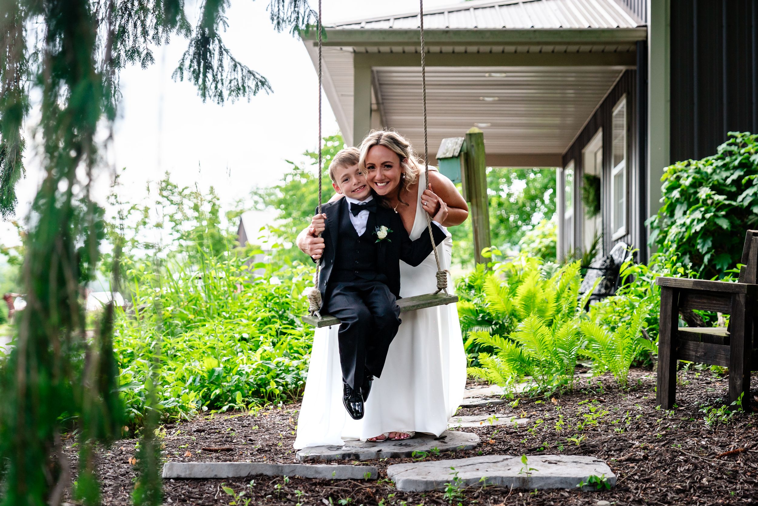 A bride in a white dress hugs a young boy in a tuxedo outside a venue.