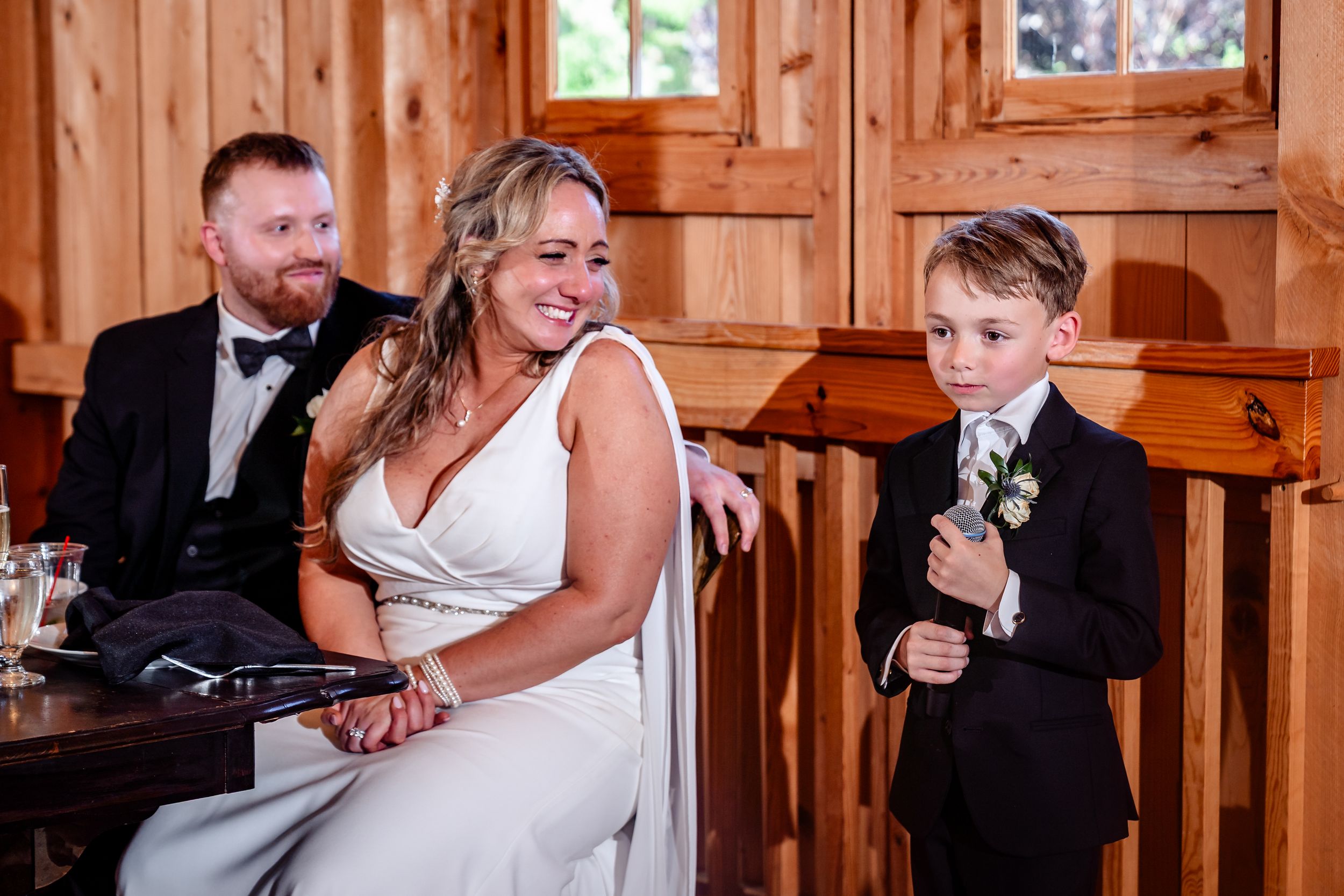 Bride in white gown sits with groom in black suit and young ring bearer at wedding reception.