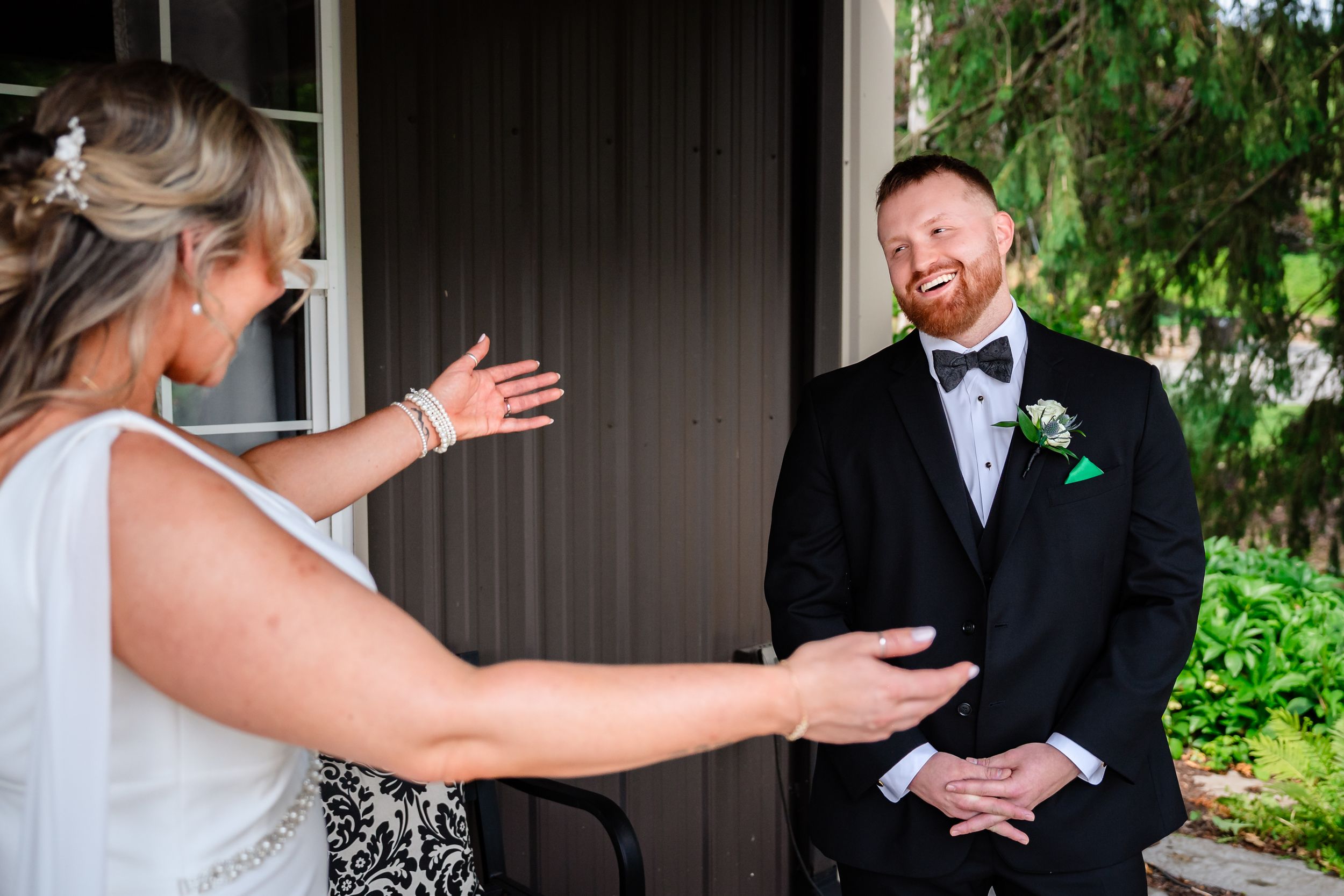 Joyful groom in black tuxedo reacting with delight during first look moment with bride outdoors.