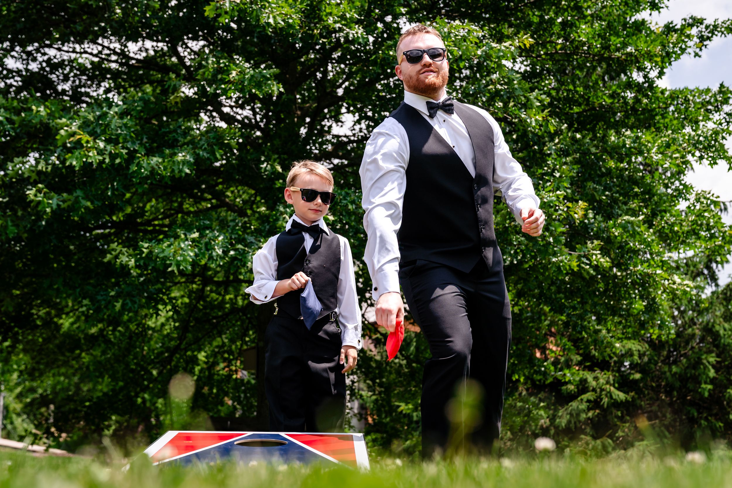 Groom and young ring bearer in matching black vests and sunglasses play cornhole outdoors on a sunny wedding day.