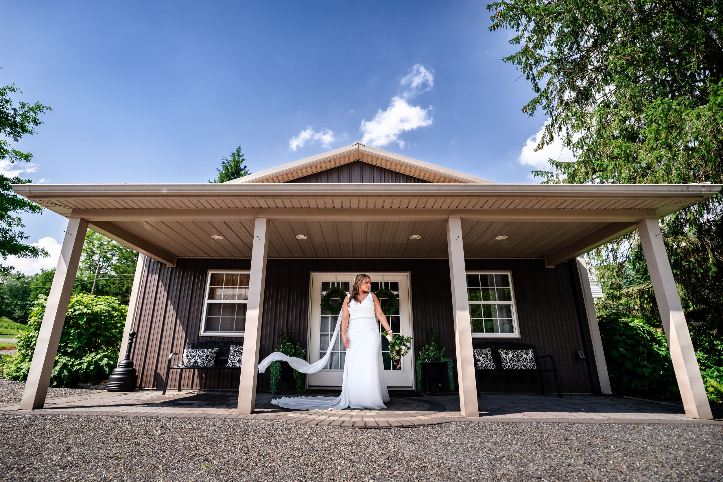 Bride in white wedding gown poses in front of a charming gray venue building on a sunny day.
