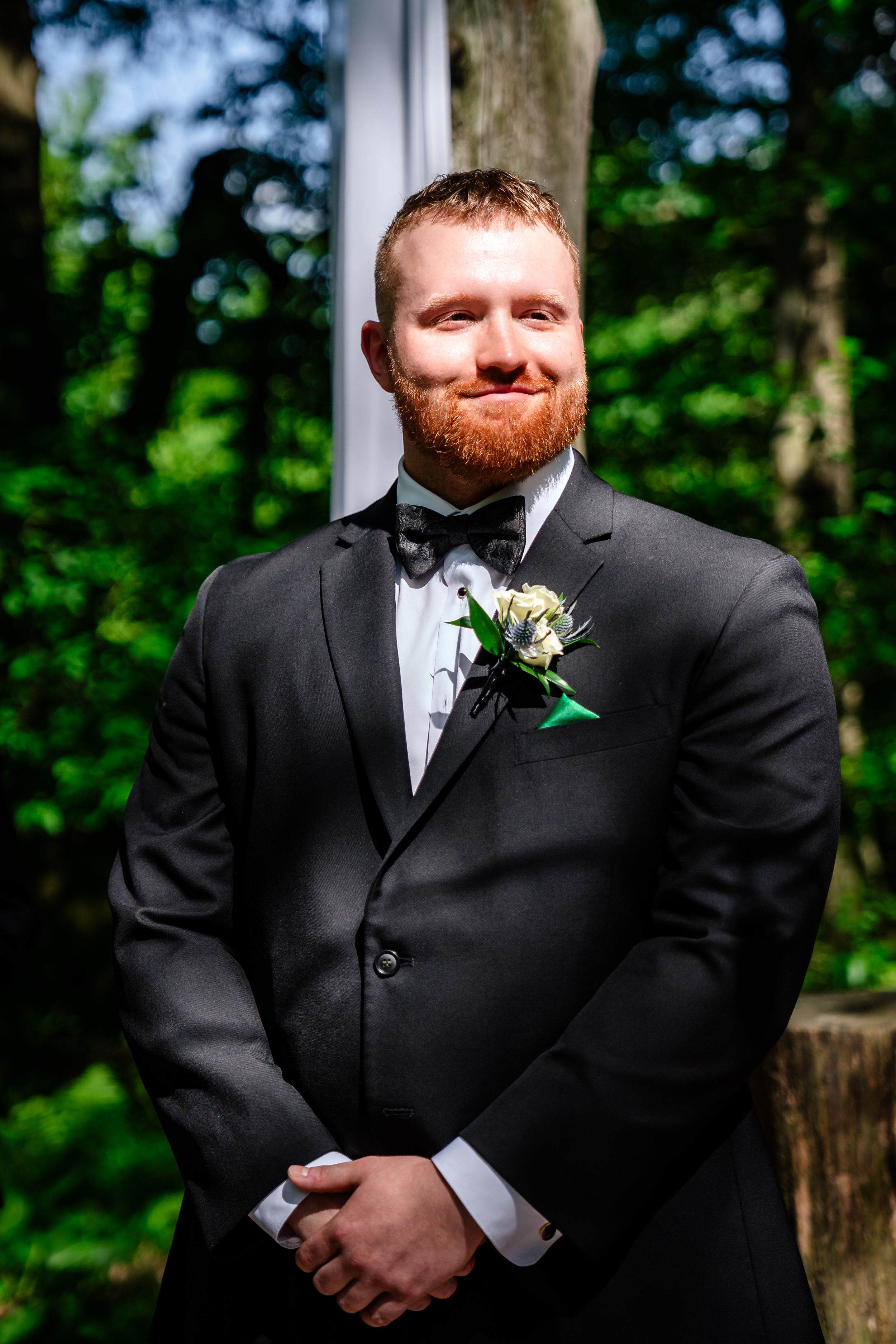 Smiling groom with red beard wearing black tuxedo and boutonniere stands outdoors on wedding day.