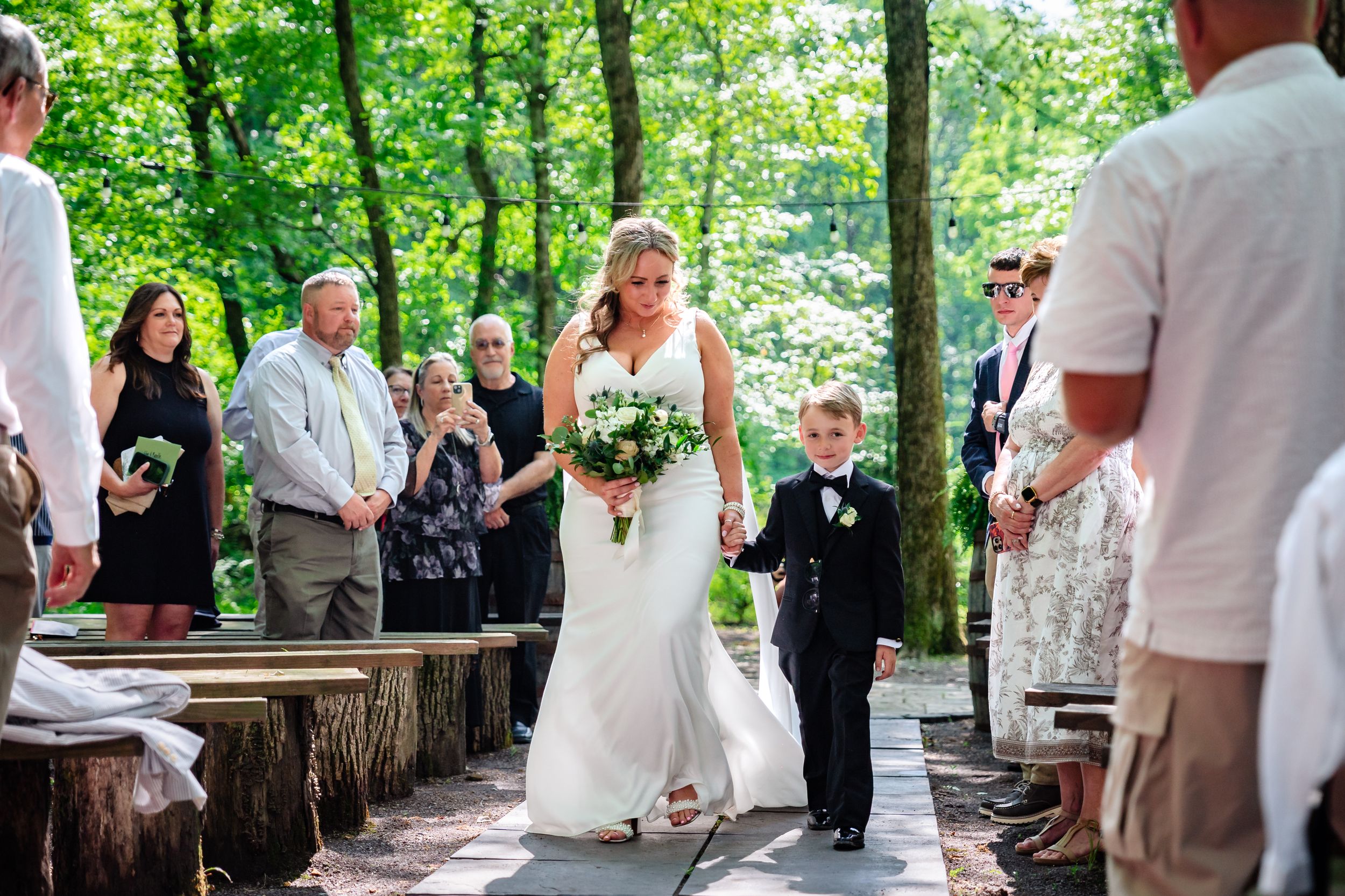 Bride in white fitted gown walks down outdoor aisle with young ring bearer through lush green forest.