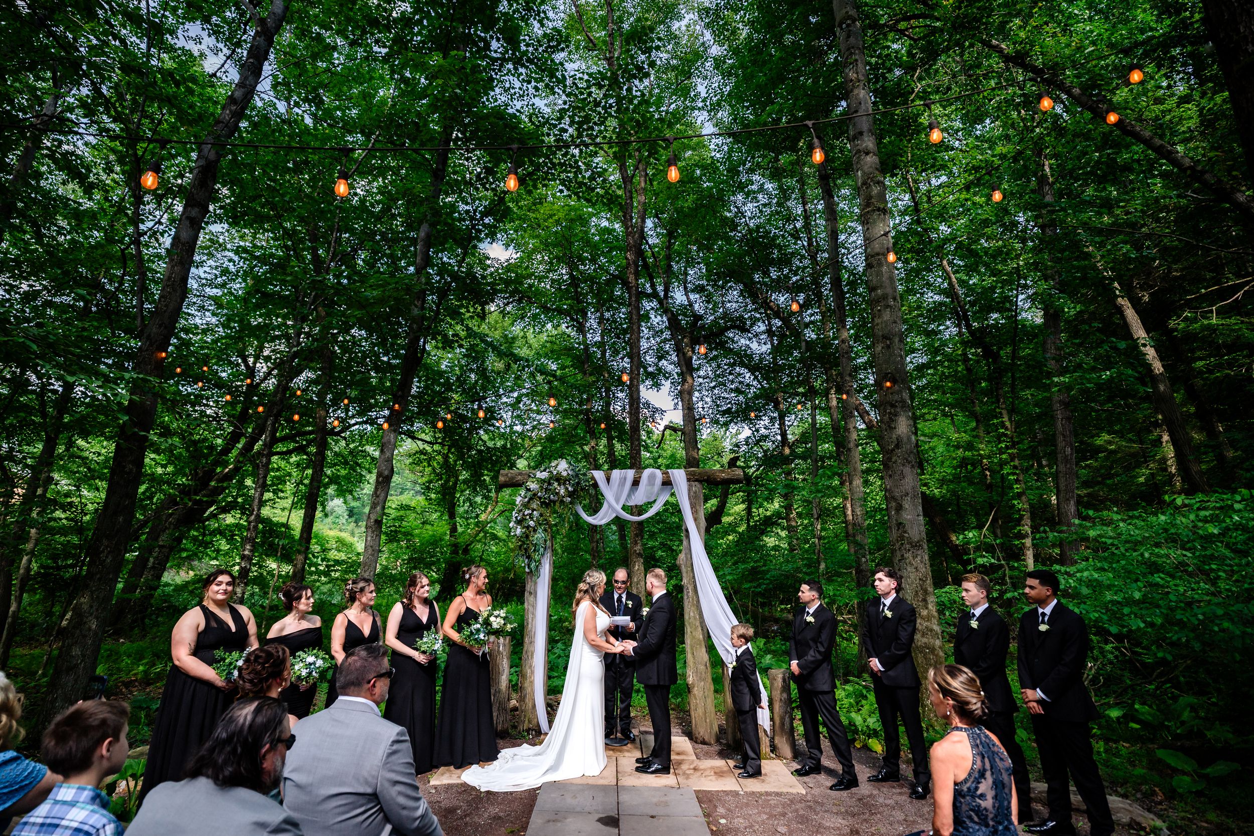 Wedding ceremony in a forest clearing with bridal party in black under a draped arch with string lights.