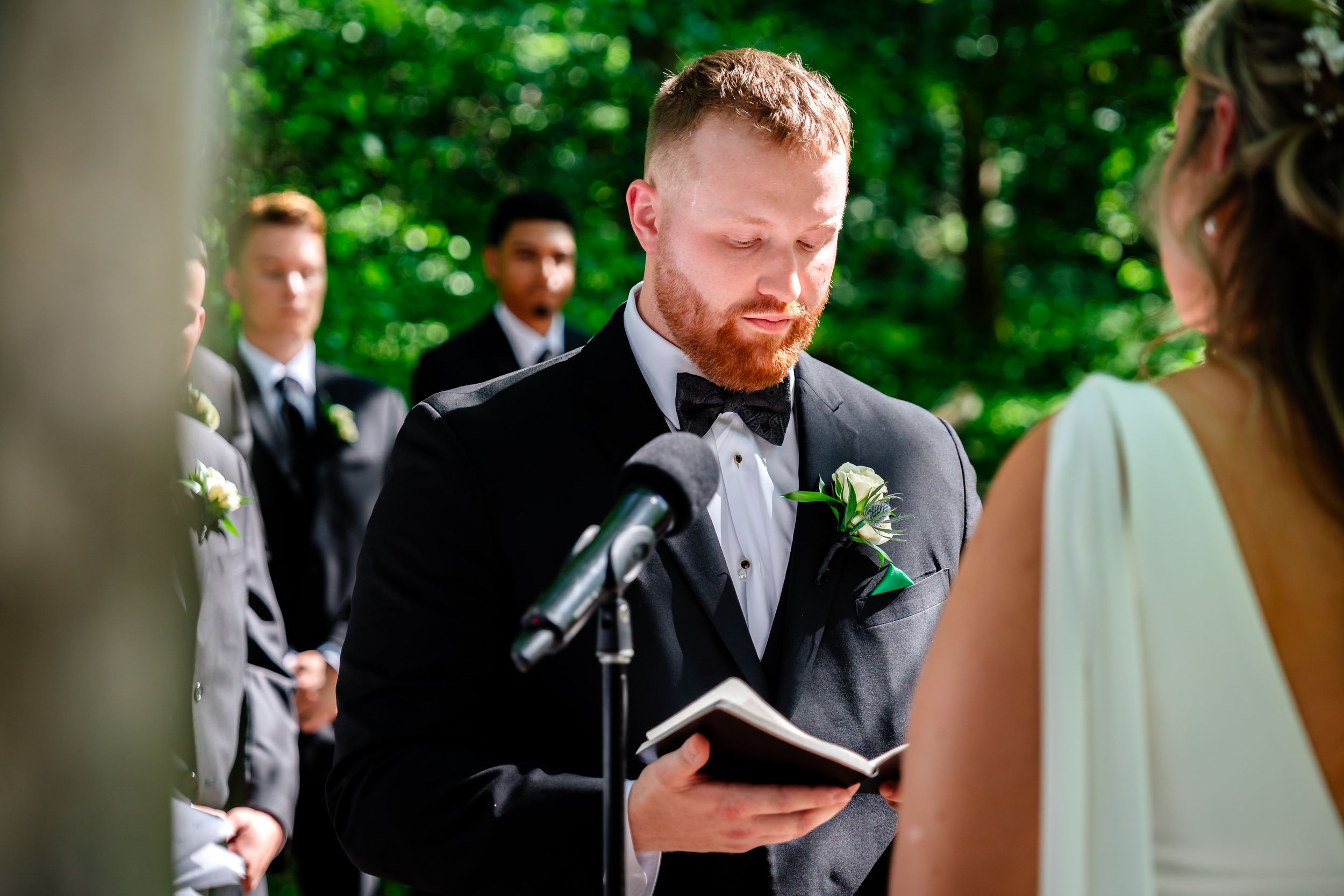 Groom with red beard reading vows from a book at outdoor wedding ceremony with groomsmen in background.