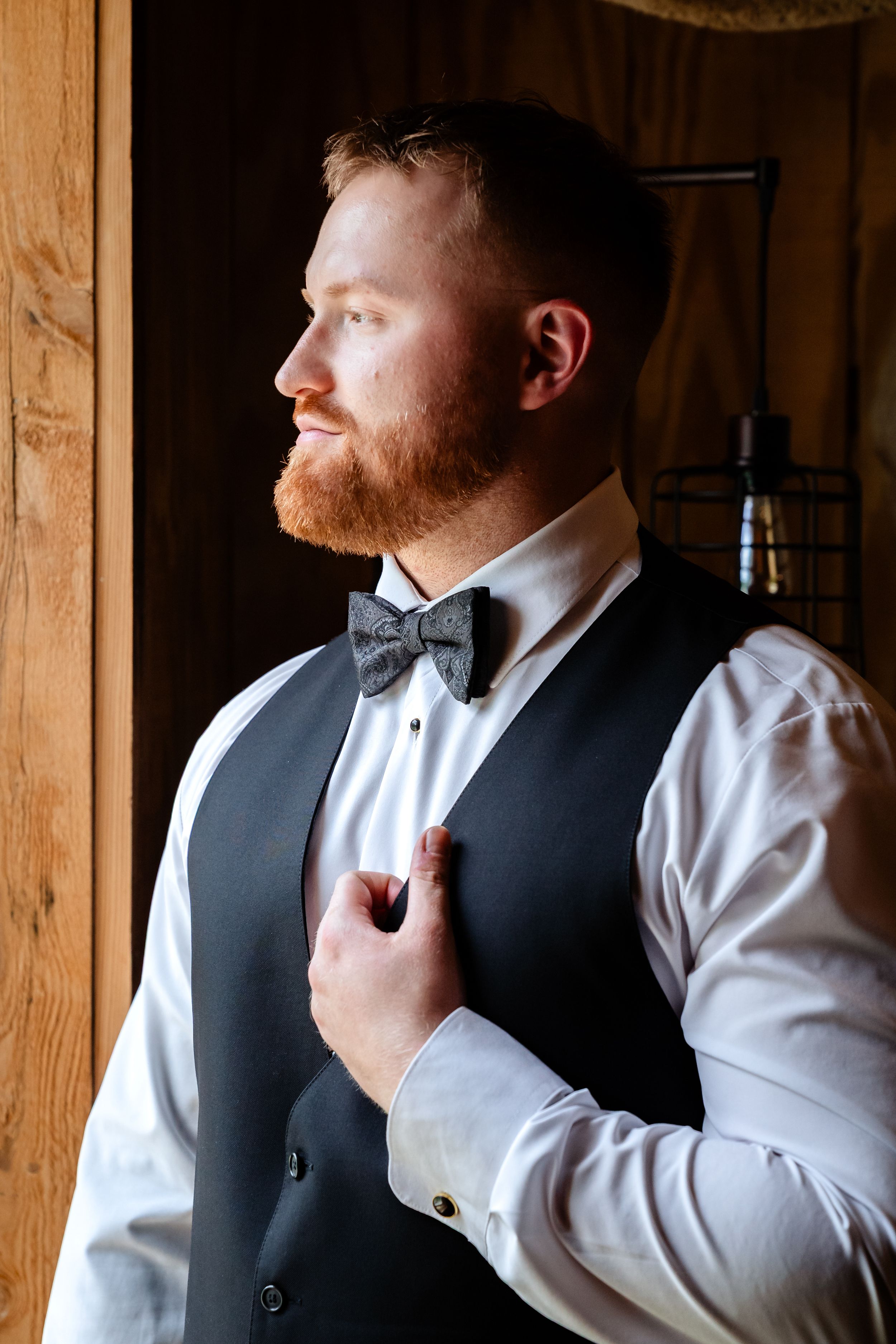 Dapper groom in white shirt, gray vest and bow tie adjusting his lapel while gazing out a window.