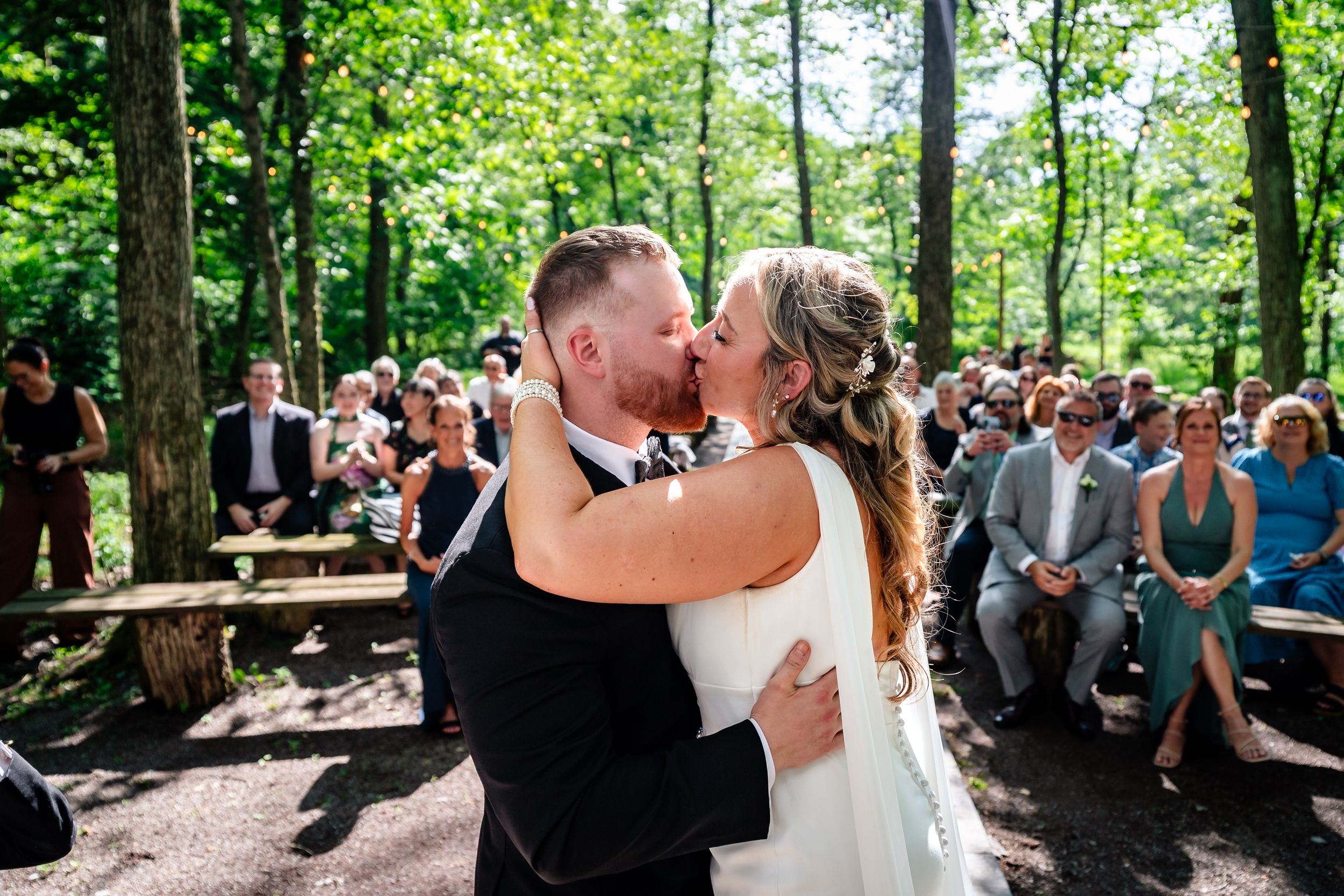 Bride and groom share first kiss at outdoor forest wedding ceremony with guests.