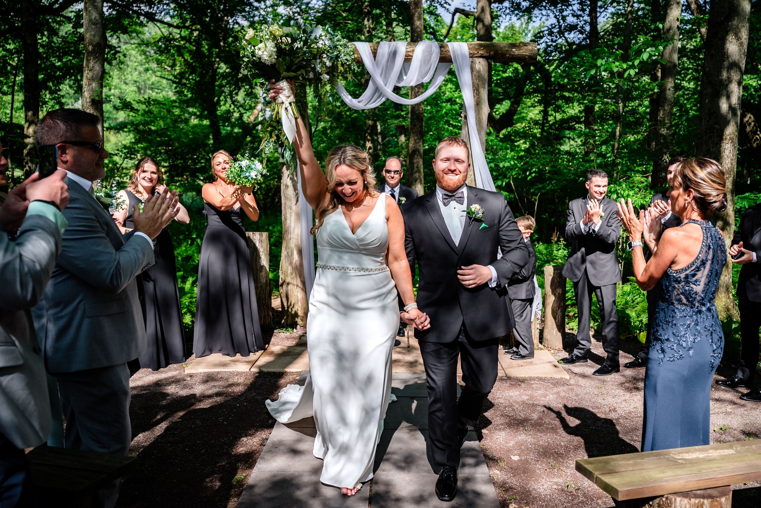 Bride and groom exit outdoor woodland wedding ceremony as guests celebrate.