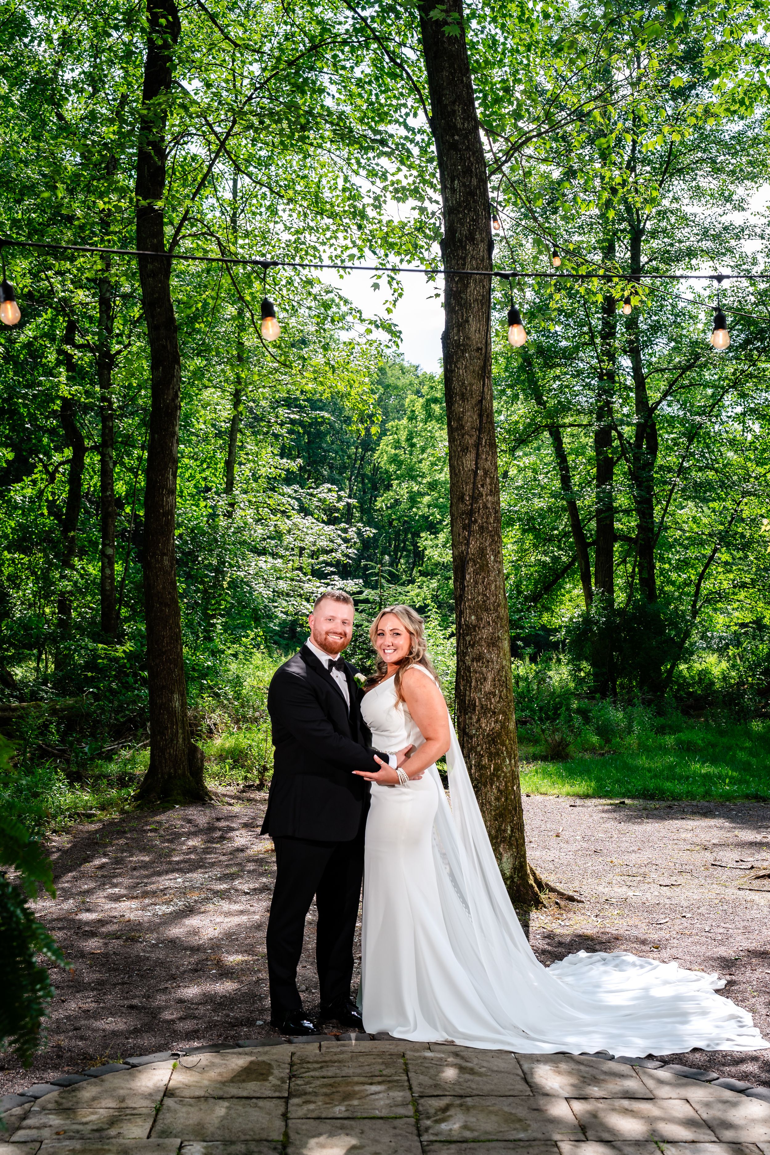 A bride and groom pose together in a lush green forest setting, with string lights overhead on their wedding day.