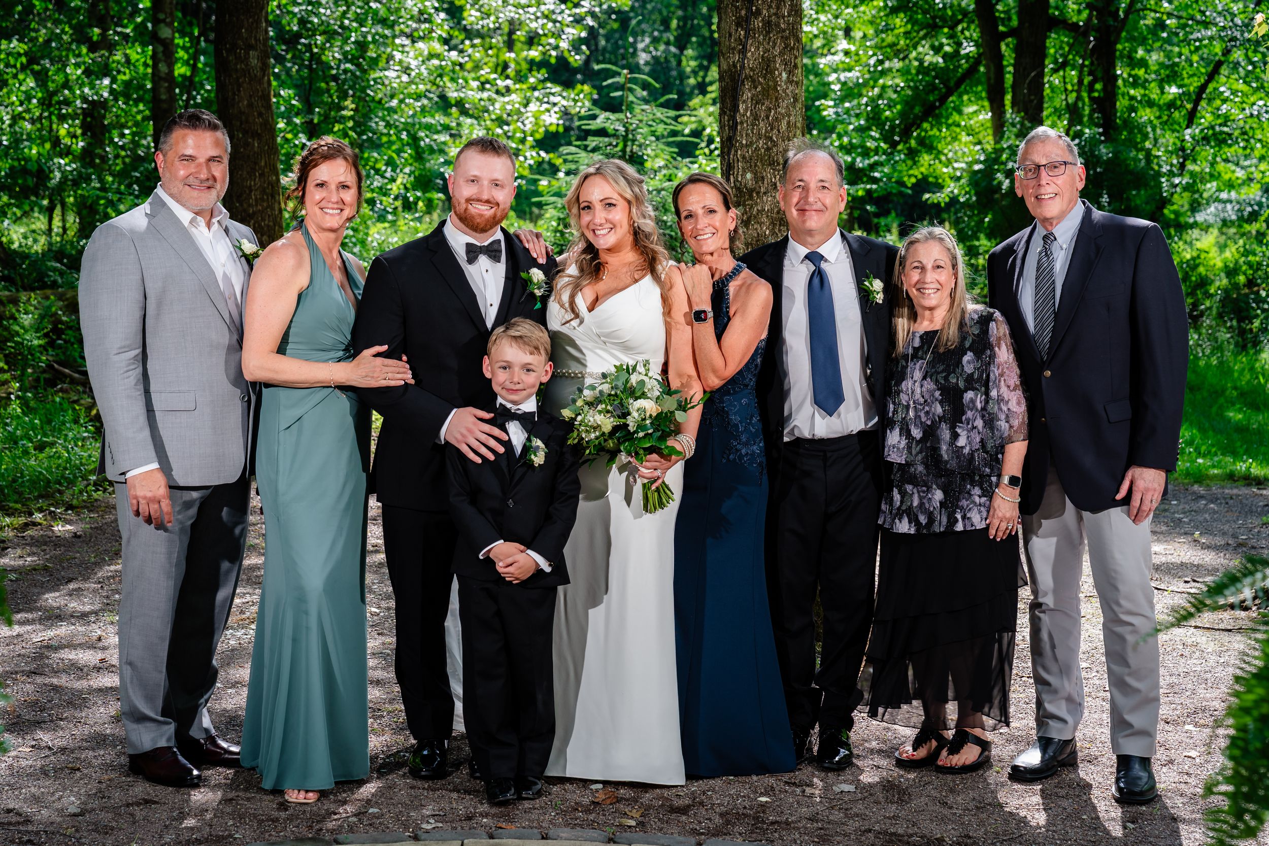 Wedding party of eight adults and a young boy posing outdoors among lush green trees.