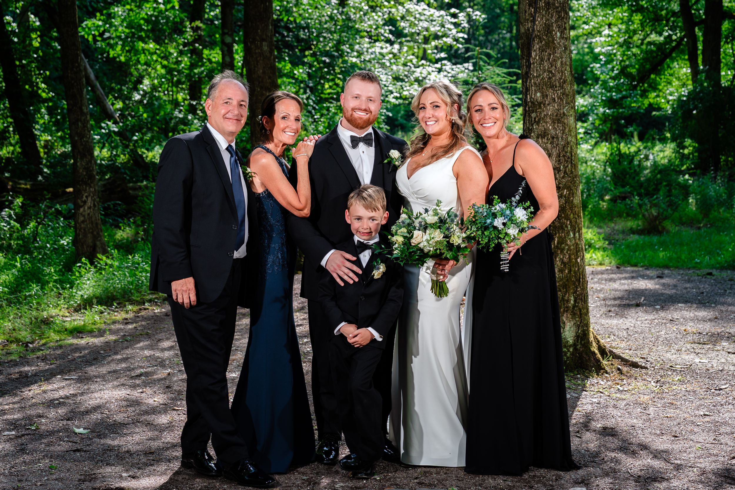 Family of six including bride, groom, and young ring bearer posing outdoors near trees.