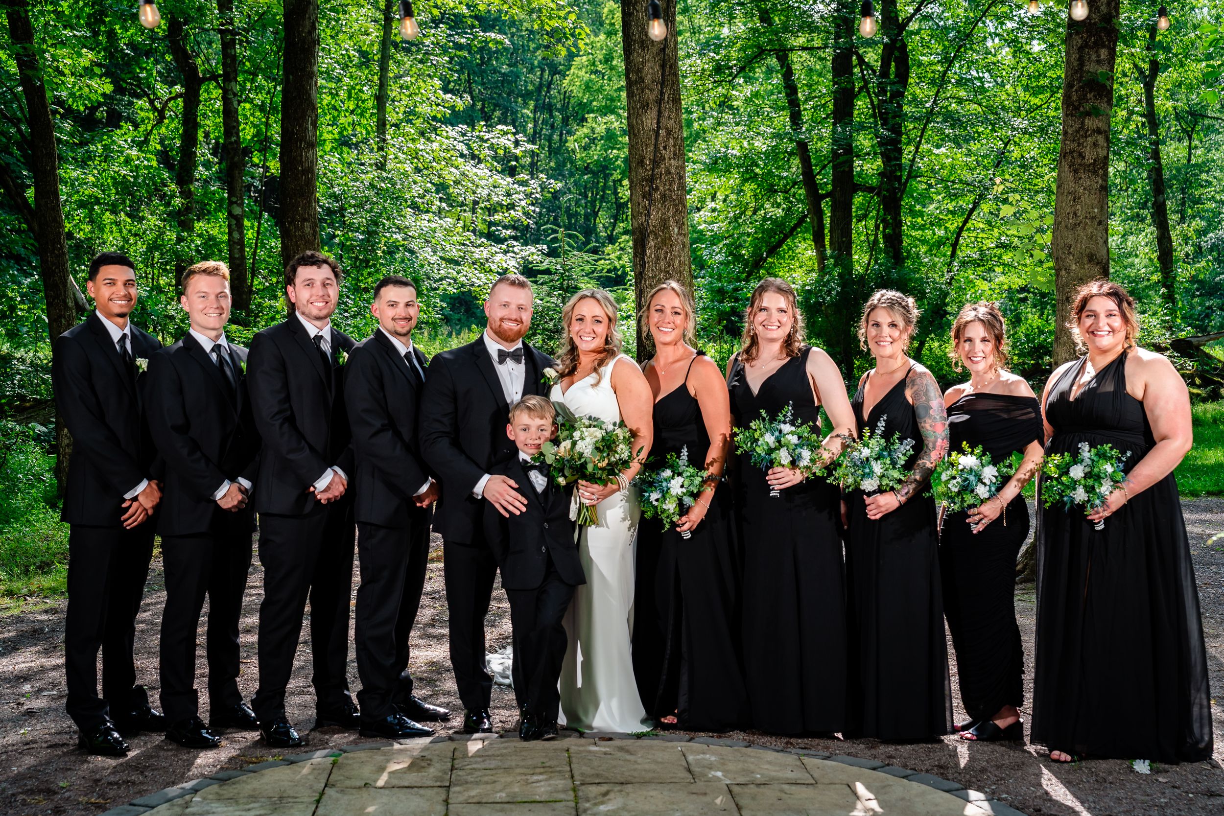 Wedding party in black attire poses outdoors among lush green trees with bride holding white bouquet.