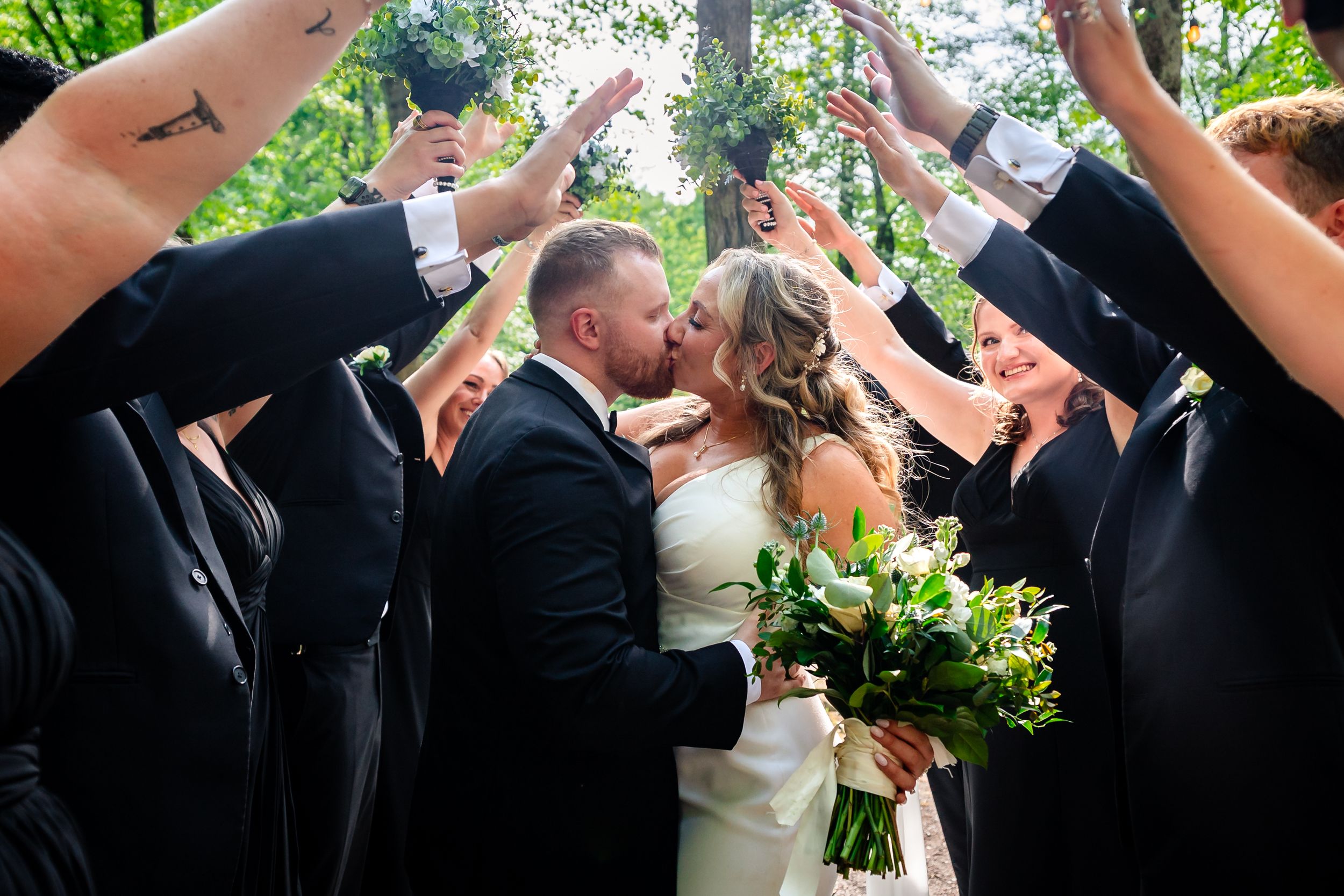 Bride and groom share a kiss under an arch of raised arms formed by wedding party members in black attire outdoors.