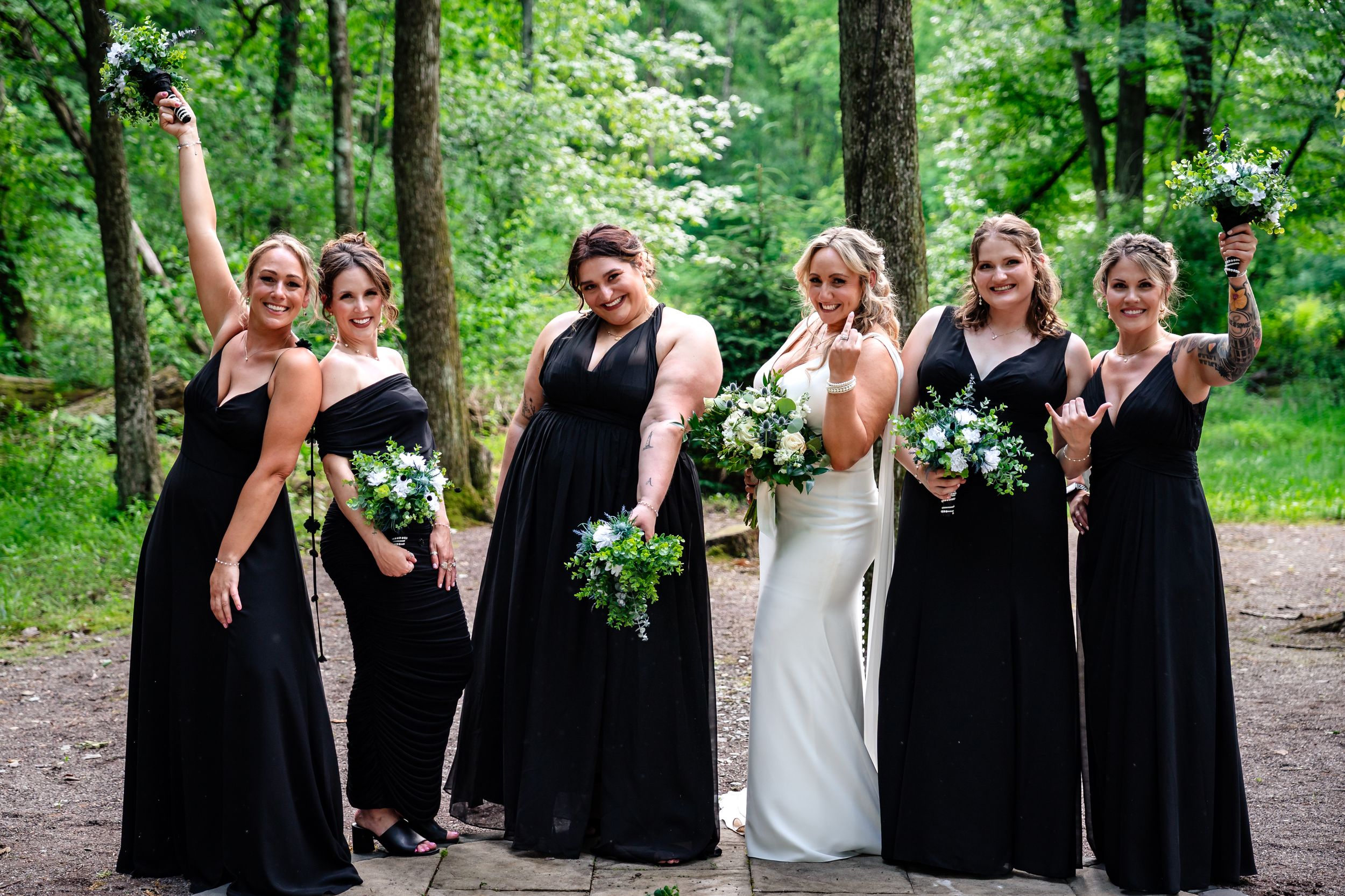 Bride poses with five bridesmaids in black gowns holding greenery bouquets in lush forest setting.