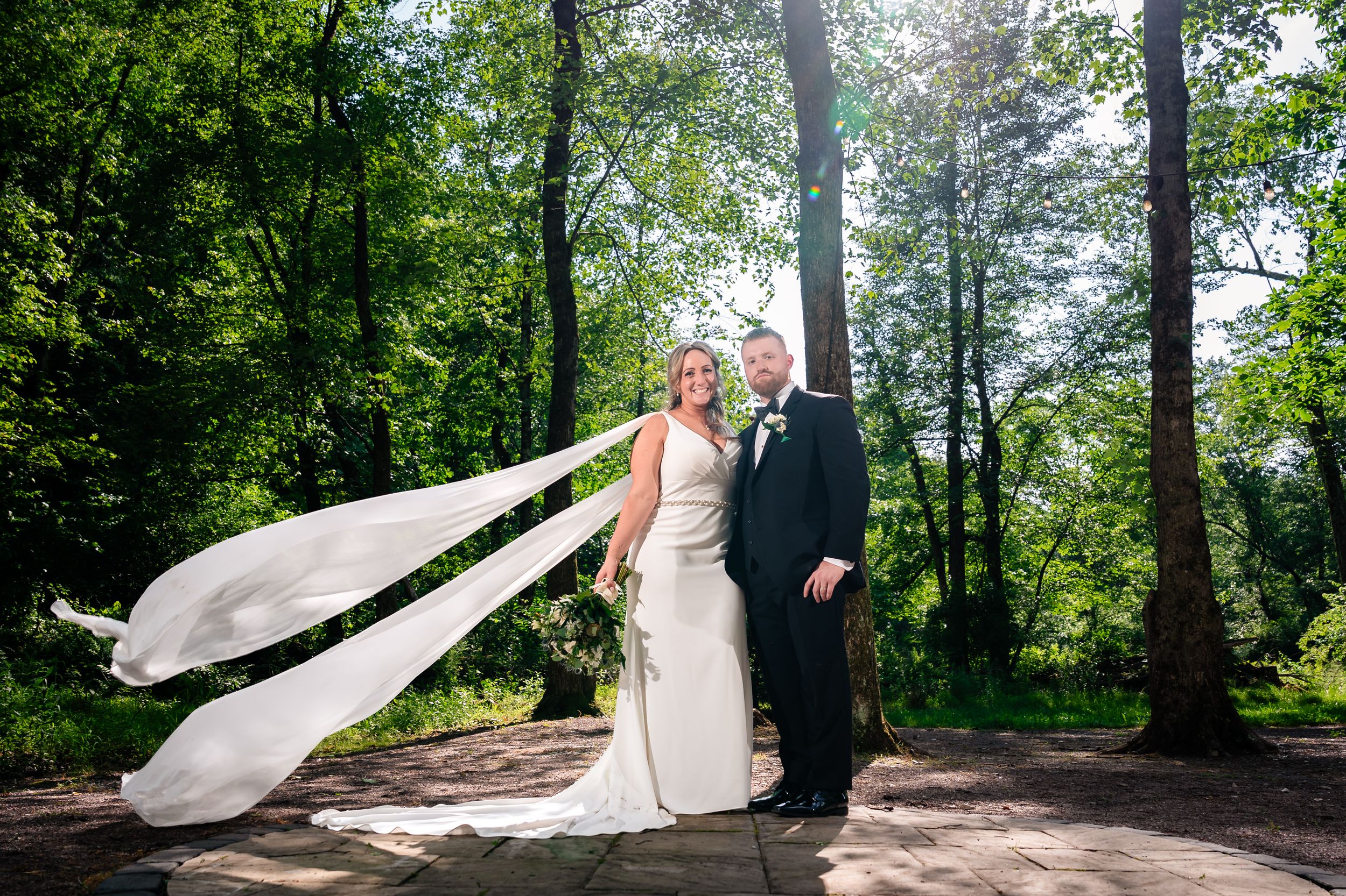 Bride and groom portrait outdoors with flowing veil caught in breeze surrounded by green trees.