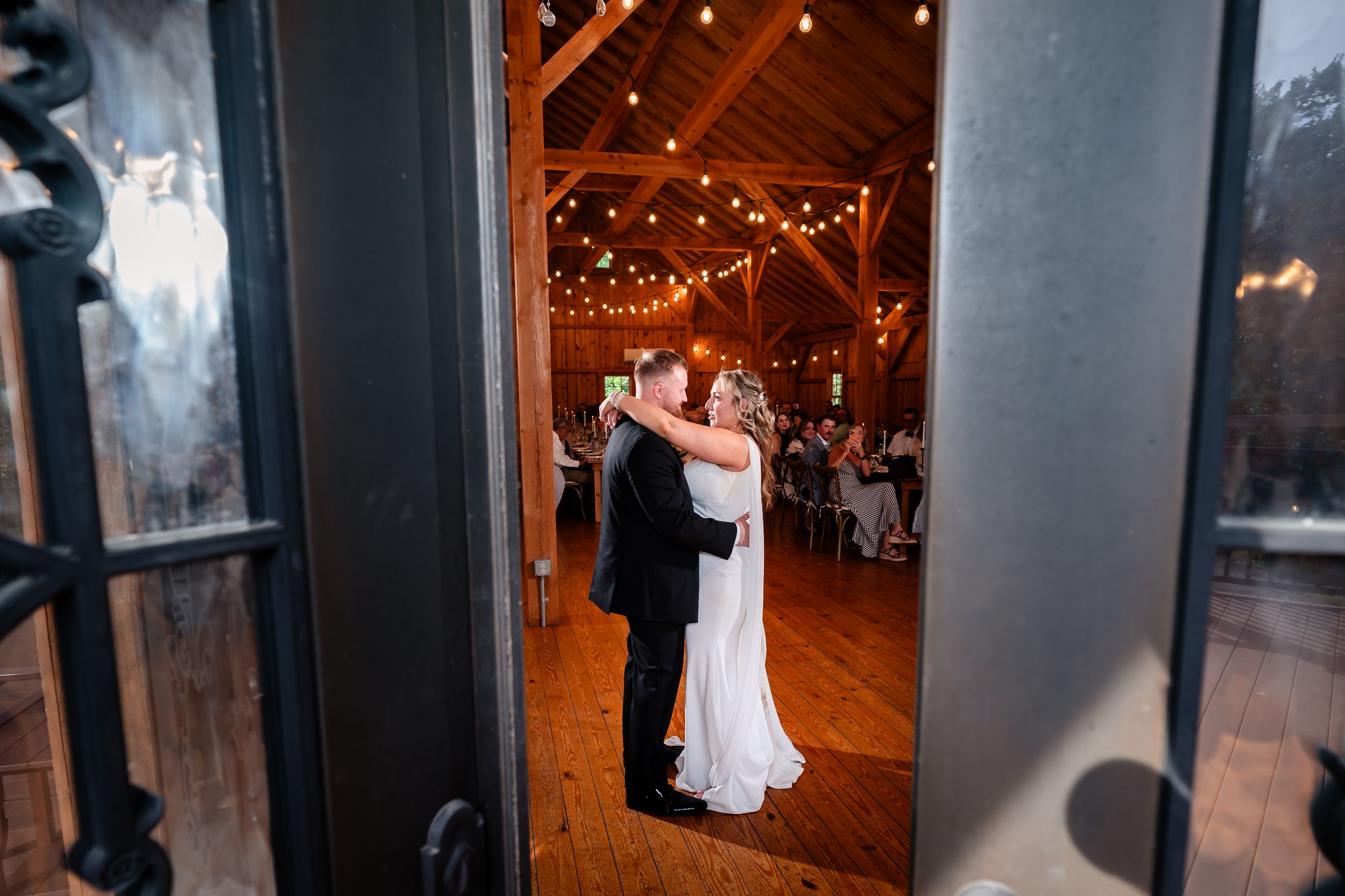 Bride and groom share first dance in rustic barn venue with warm string lights overhead.