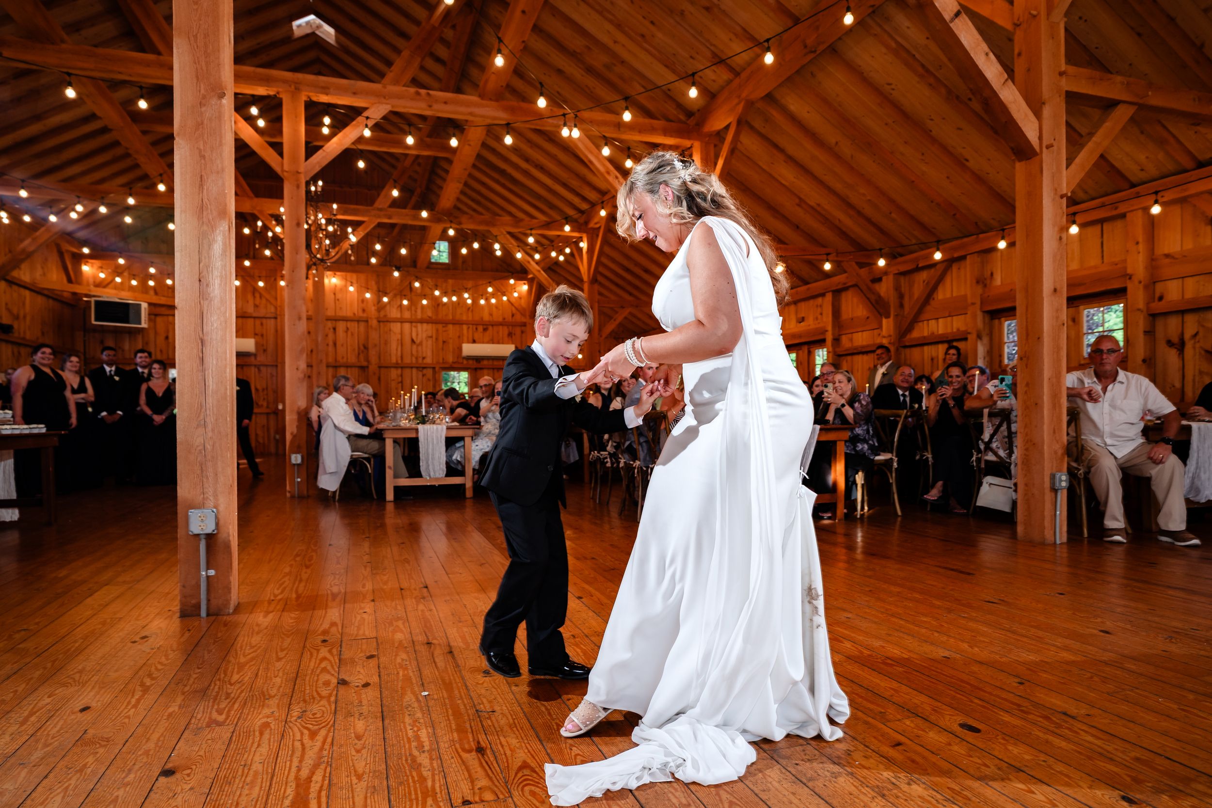 Bride in white gown dances with young boy in black suit on wooden dance floor at rustic barn wedding reception.