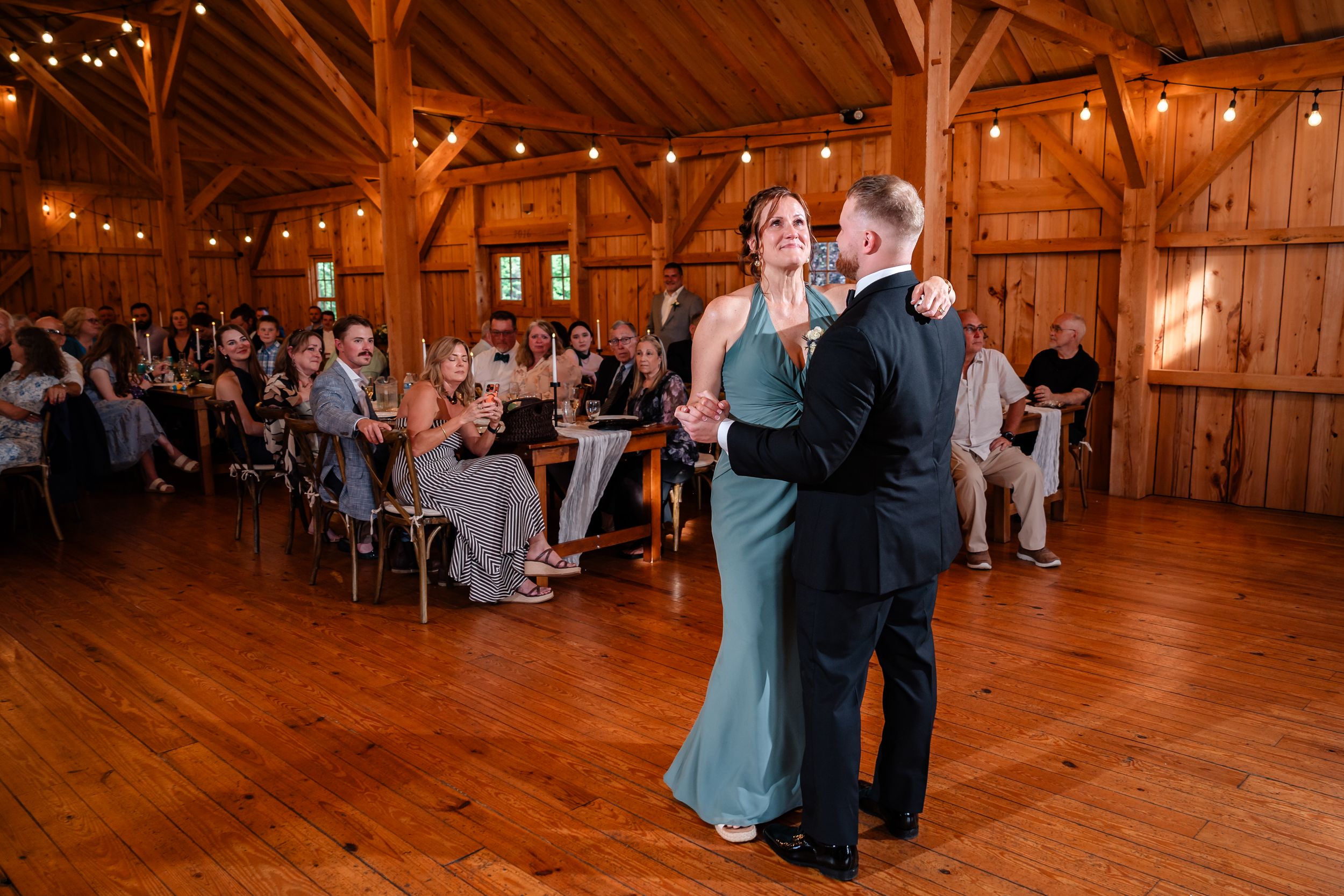 Couple shares first dance in rustic wooden barn venue with string lights and seated wedding guests.