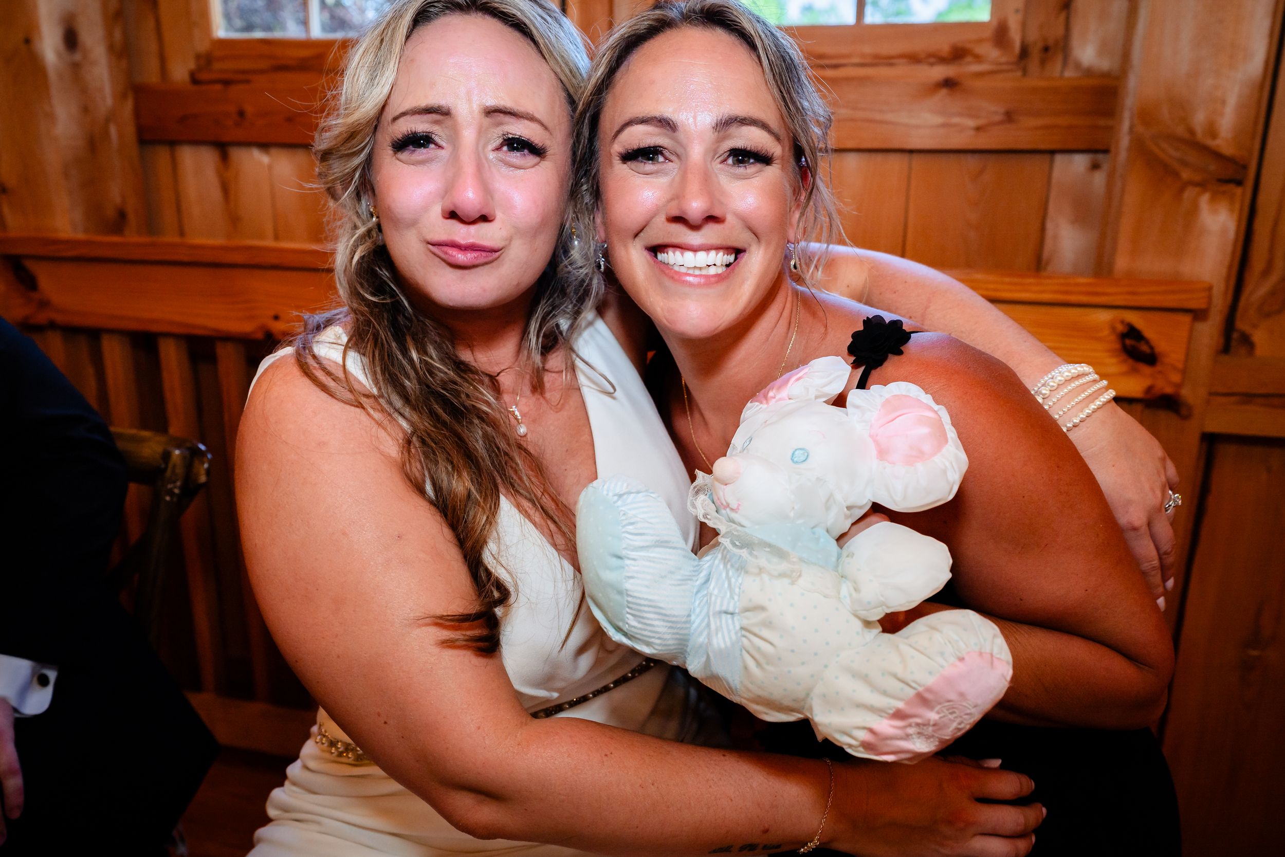 Two smiling women pose together at a wedding, one holding a stuffed animal toy inside a rustic wooden venue.