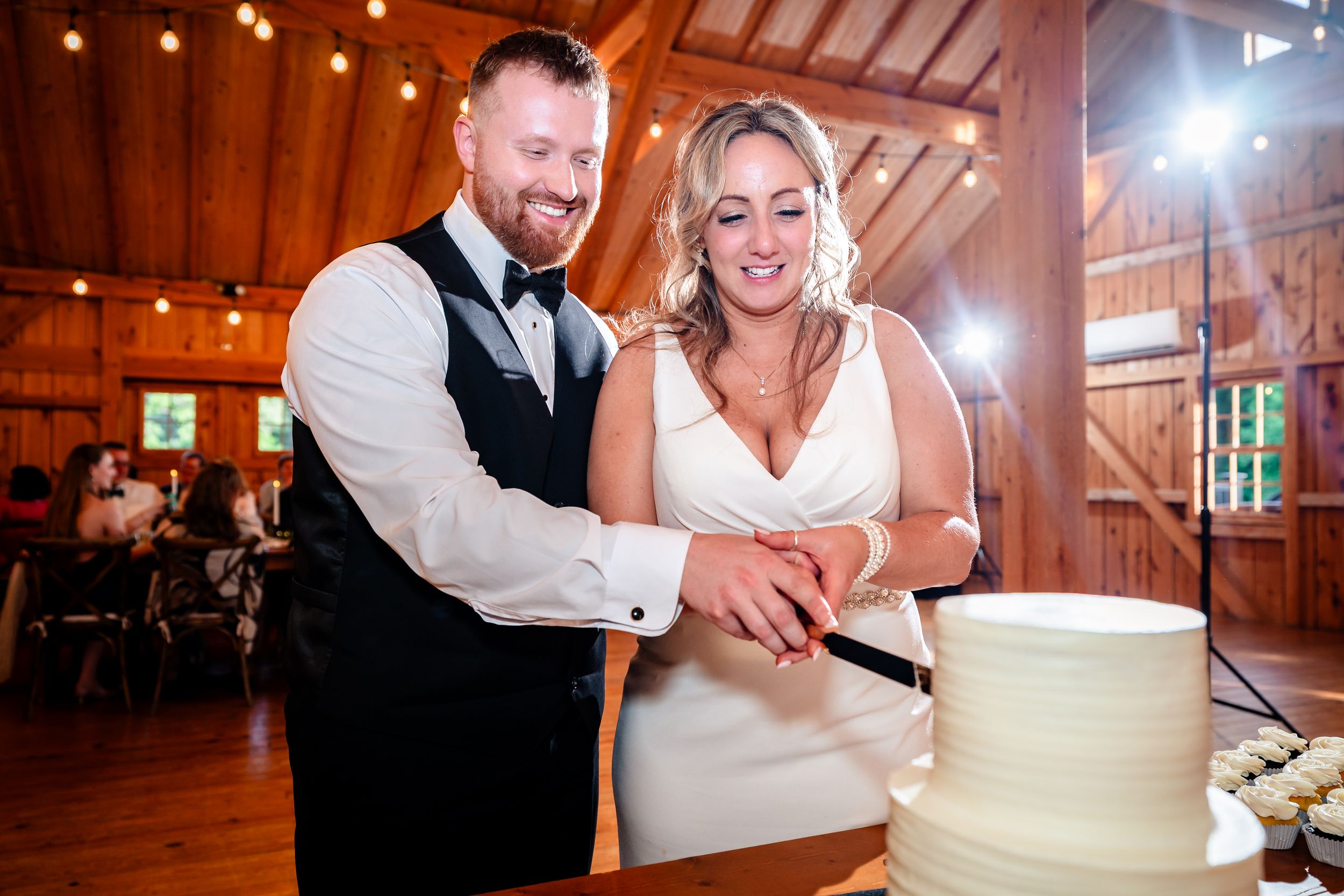 Bride and groom smiling while cutting their white wedding cake together in a rustic barn reception venue.