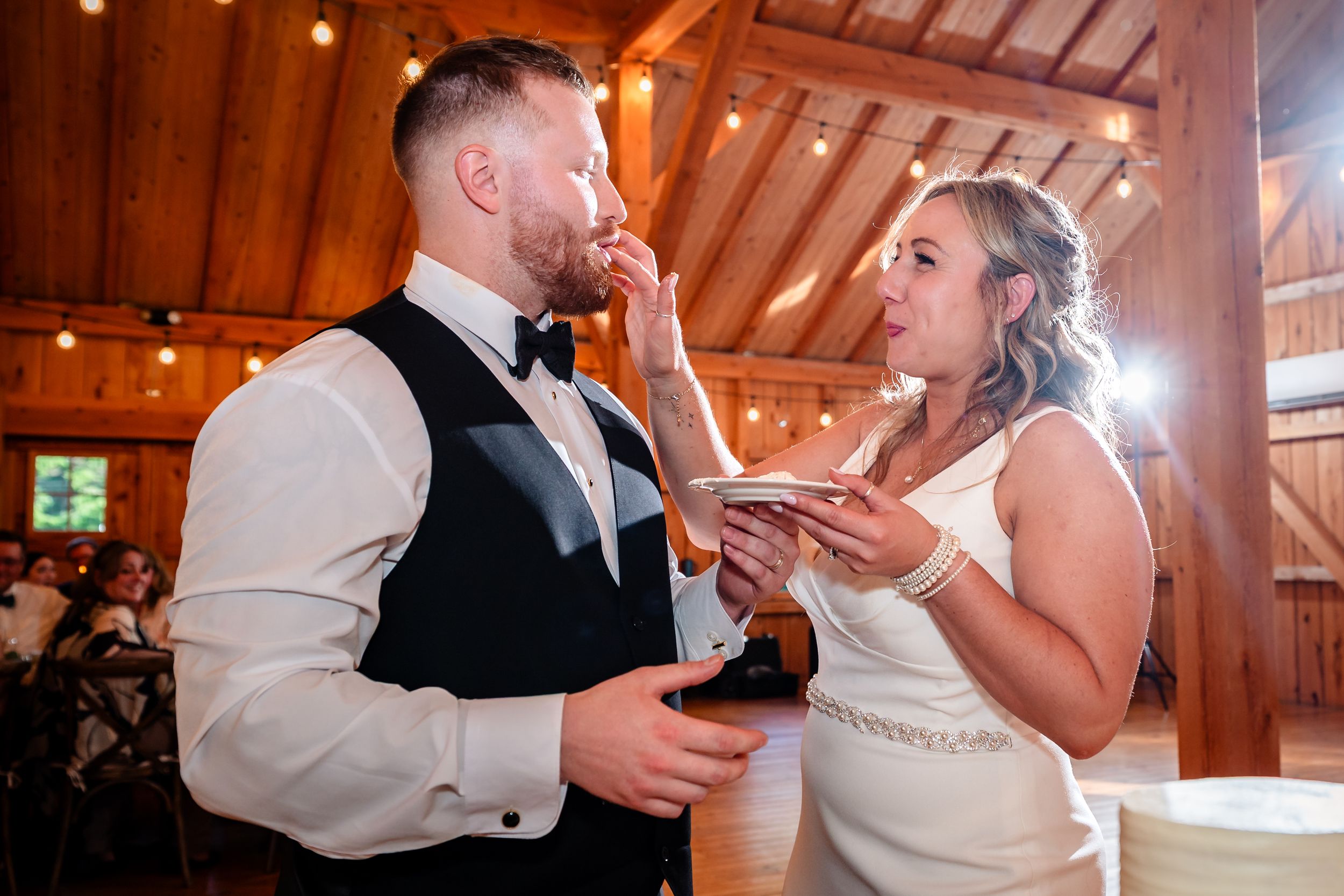 Bride feeds groom wedding cake at rustic barn reception with warm string lights.