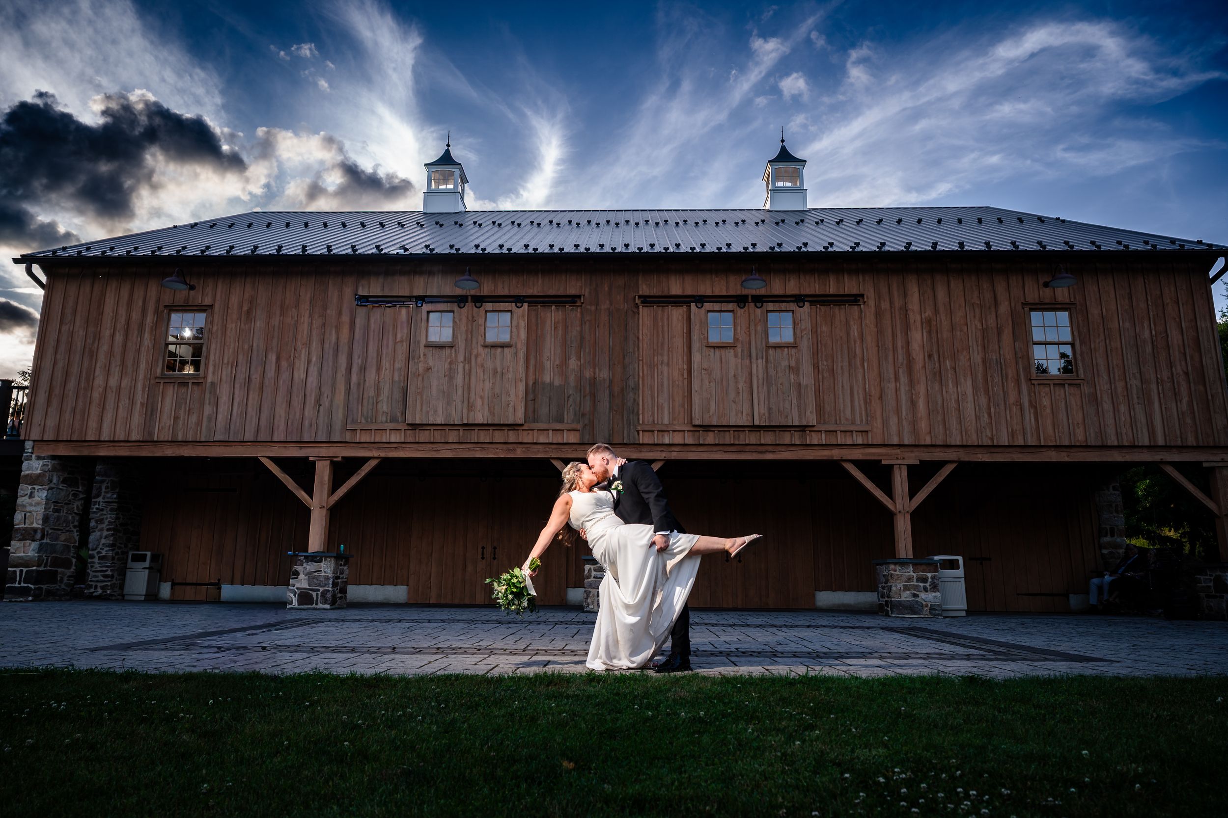 Groom dips bride in dramatic kiss outside rustic wooden barn under cloudy sky.