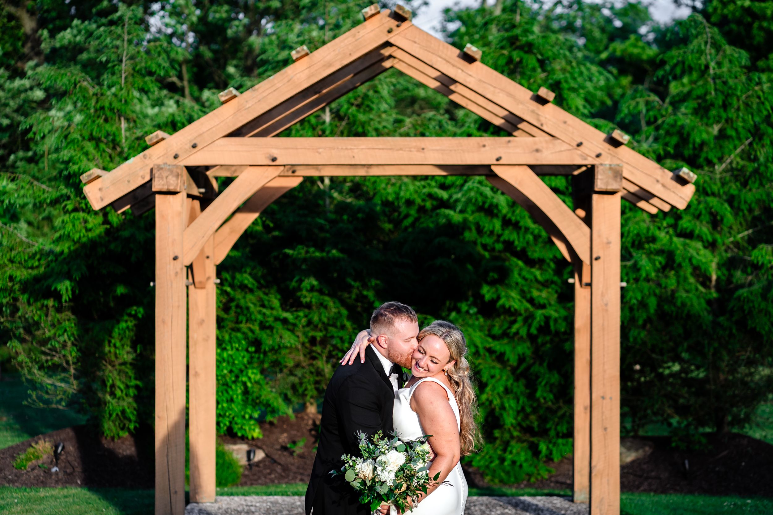 Bride and groom share a kiss under a wooden pergola surrounded by lush greenery on their wedding day.