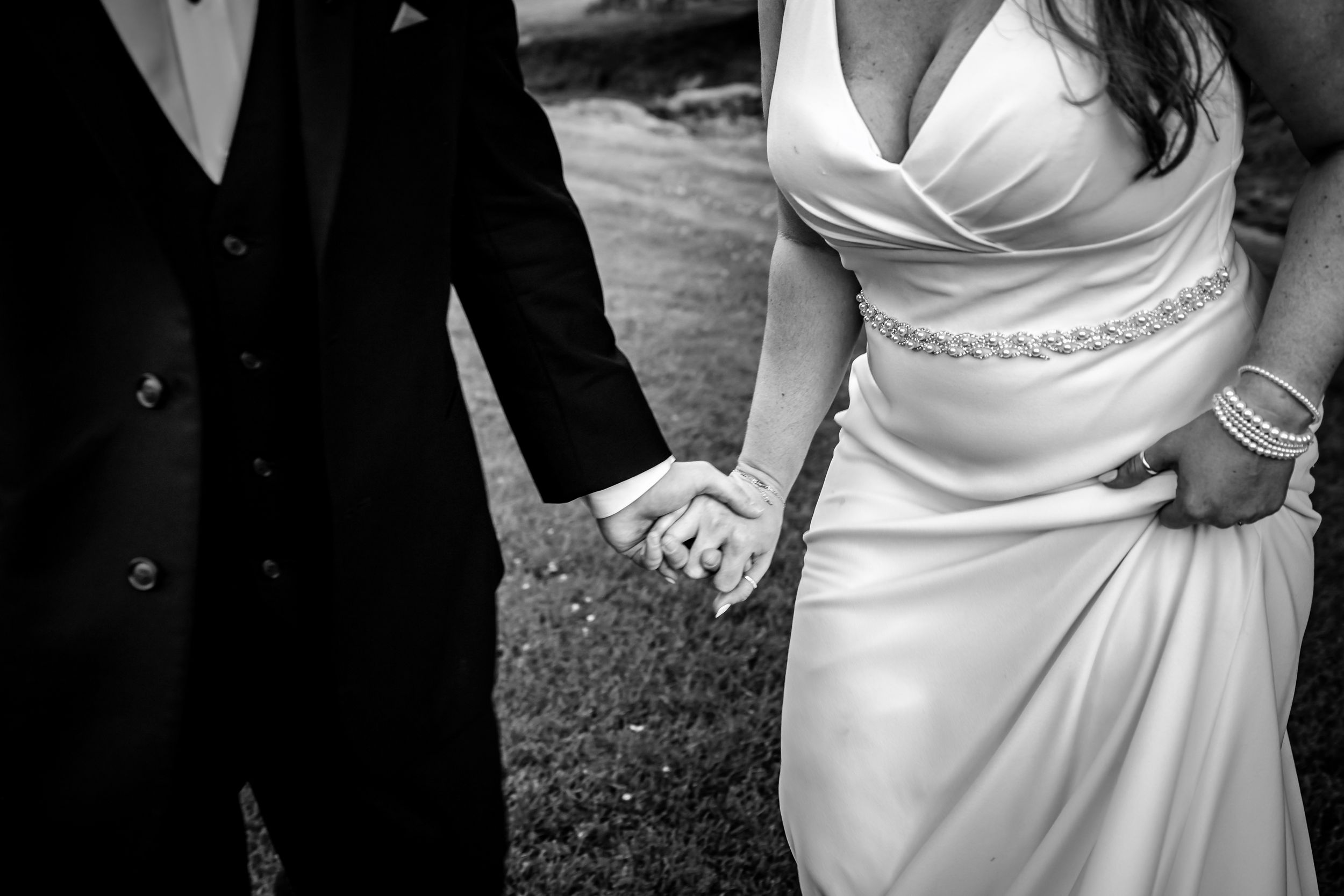 Black and white photo of bride and groom holding hands in wedding attire, showcasing elegant dress details.