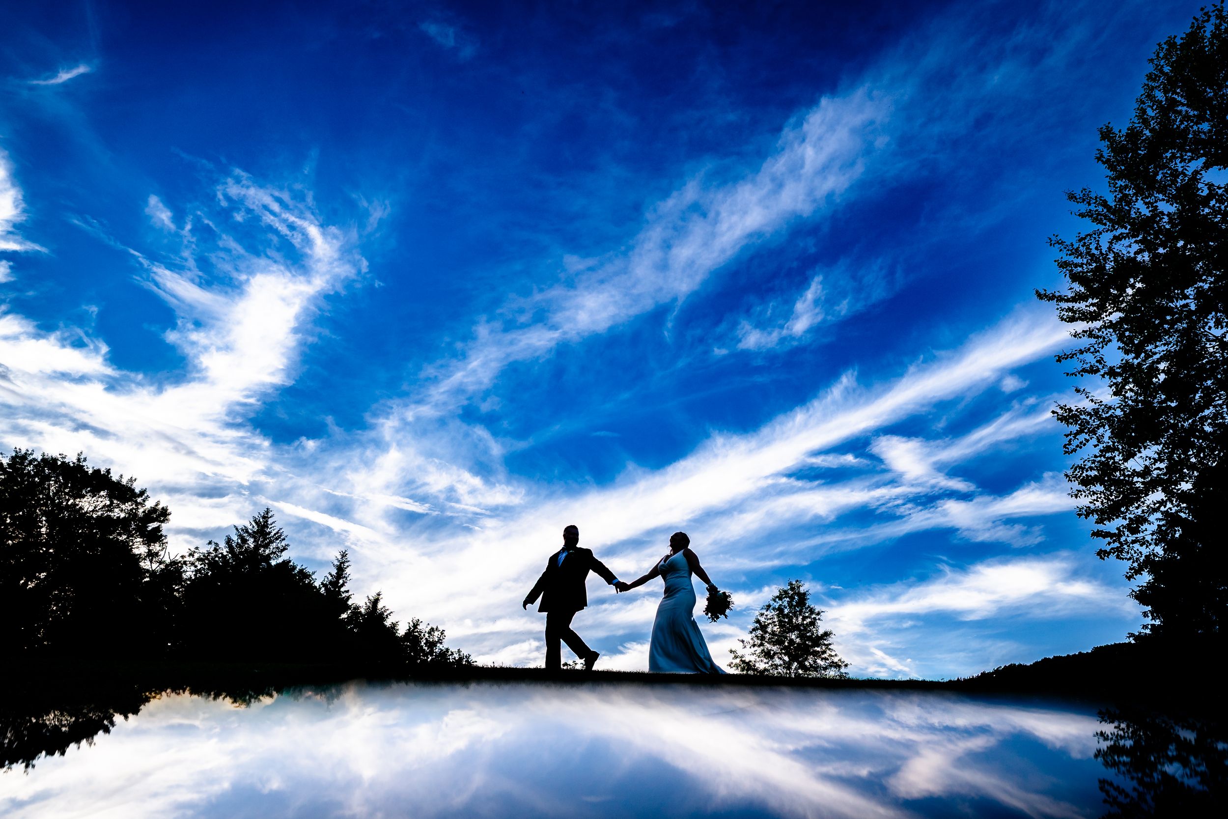 Silhouette of a bride and groom holding hands against a vivid blue sky with wispy clouds at sunset.