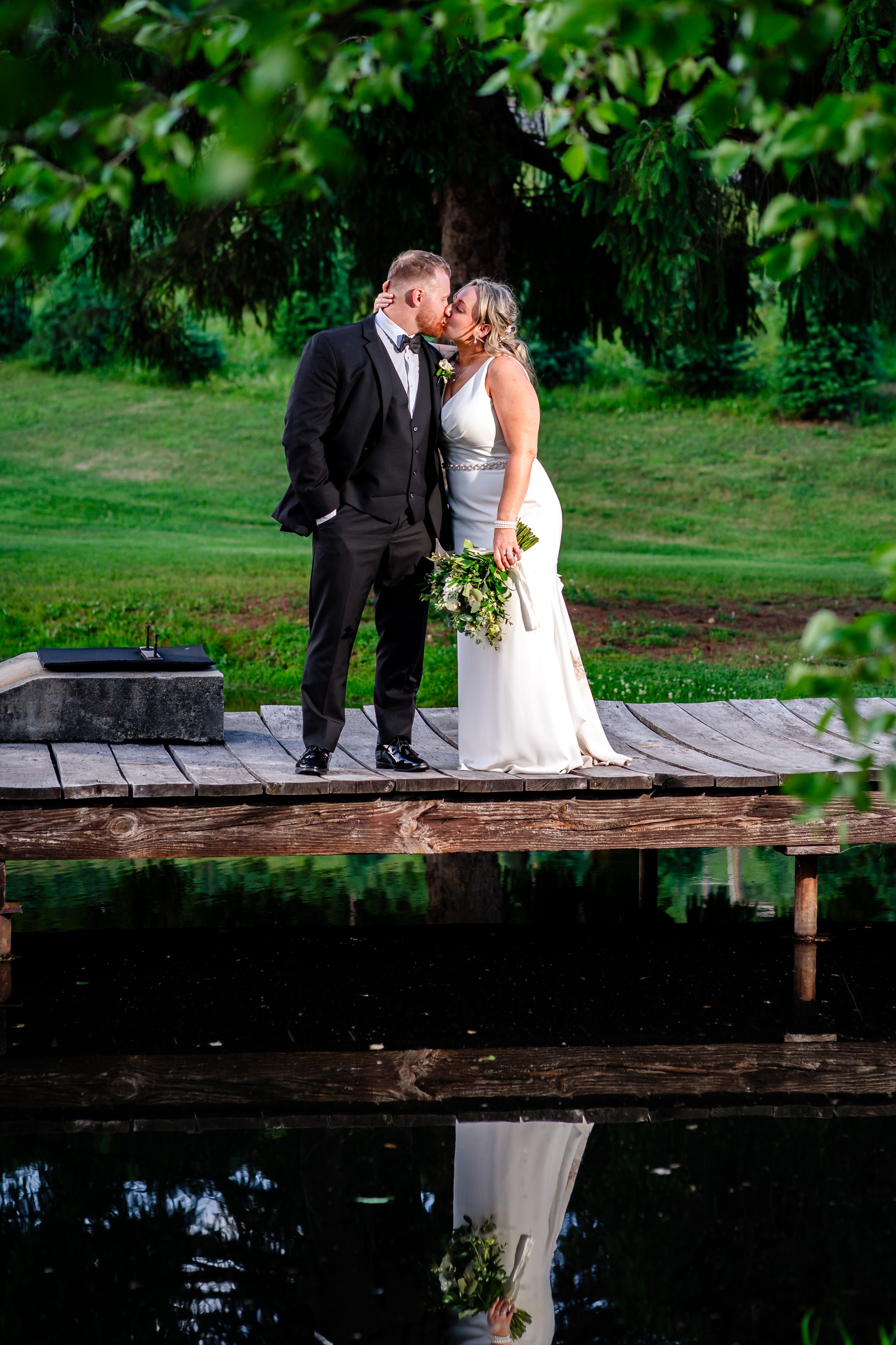 Bride and groom share a romantic kiss on a wooden dock reflected in calm water, surrounded by lush greenery.