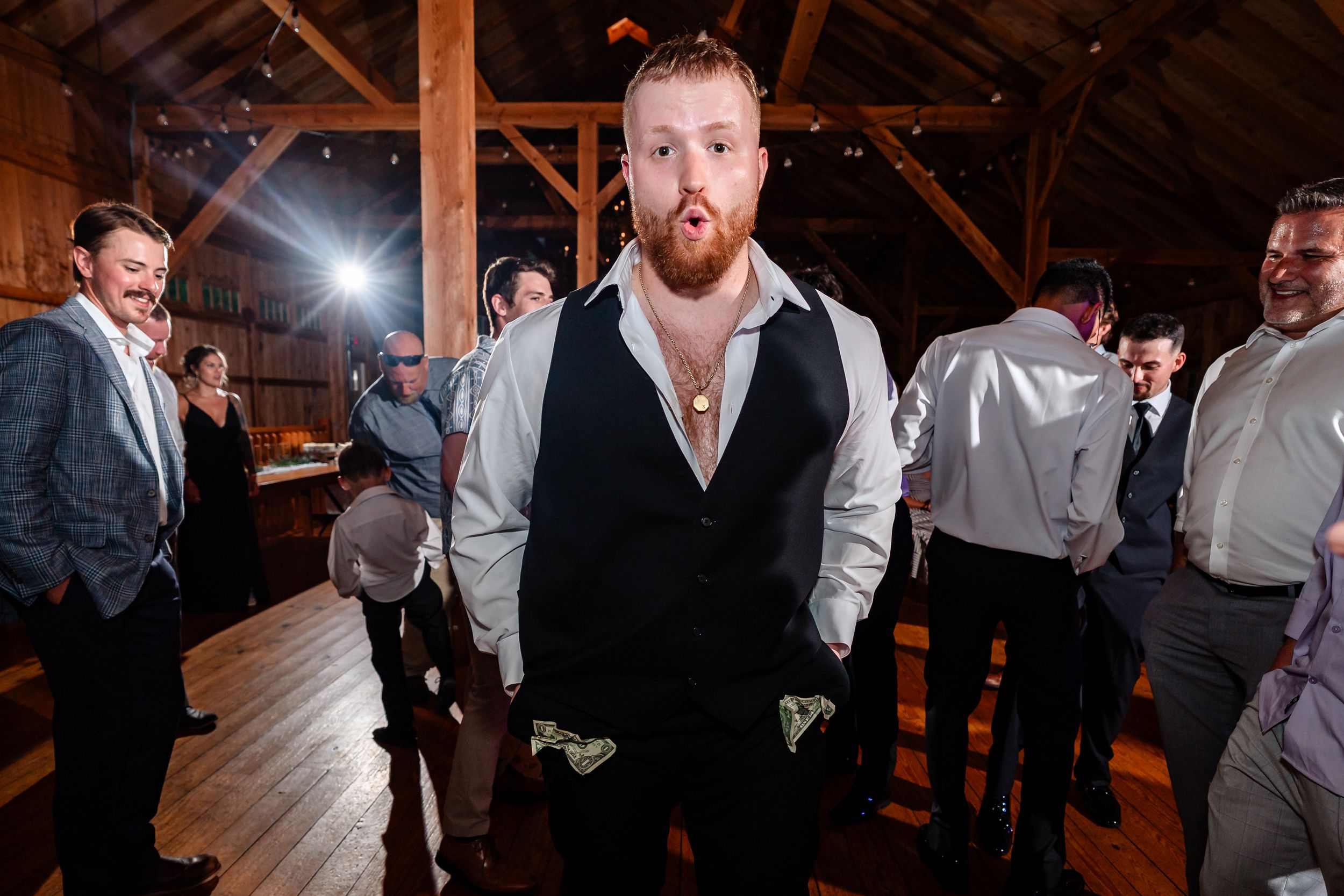 Groom in black vest and white shirt stands on dance floor at rustic barn wedding reception with guests.