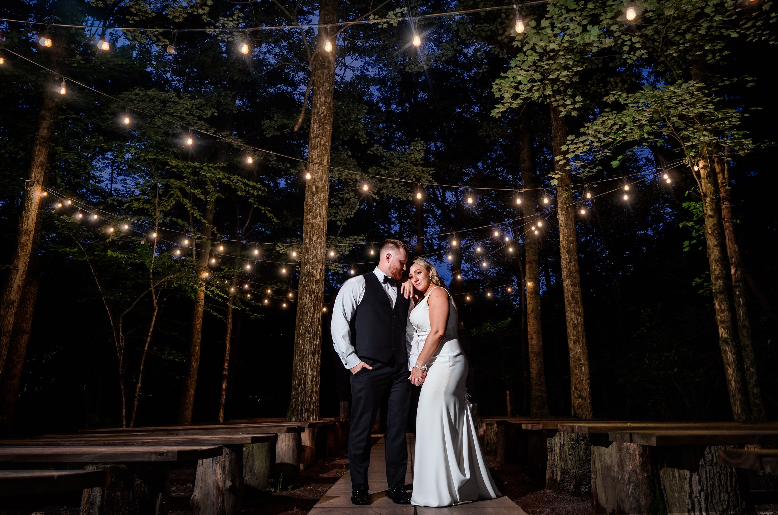 Newlywed couple shares romantic moment under string lights in wooded outdoor wedding venue at night.