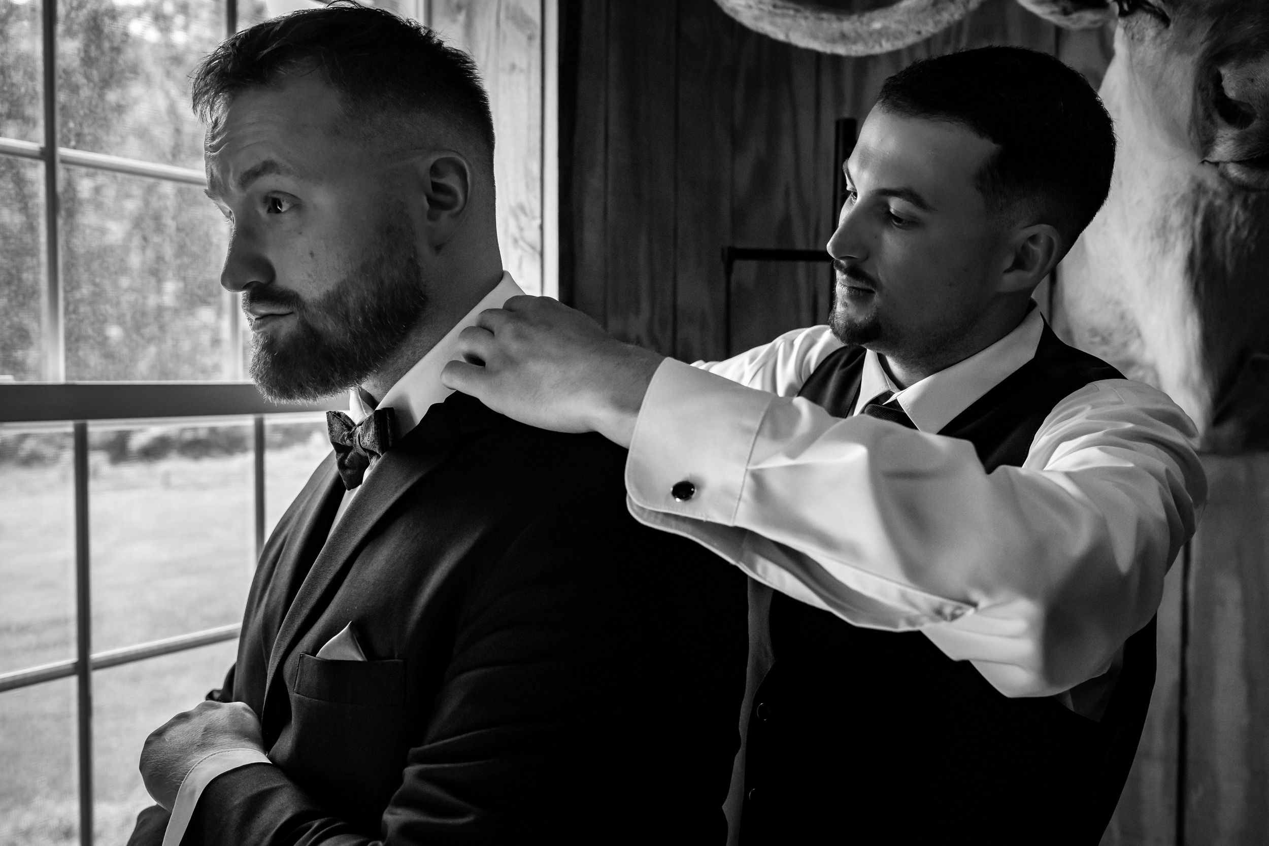 Groomsman adjusts grooms bow tie in black and white wedding preparation photo.
