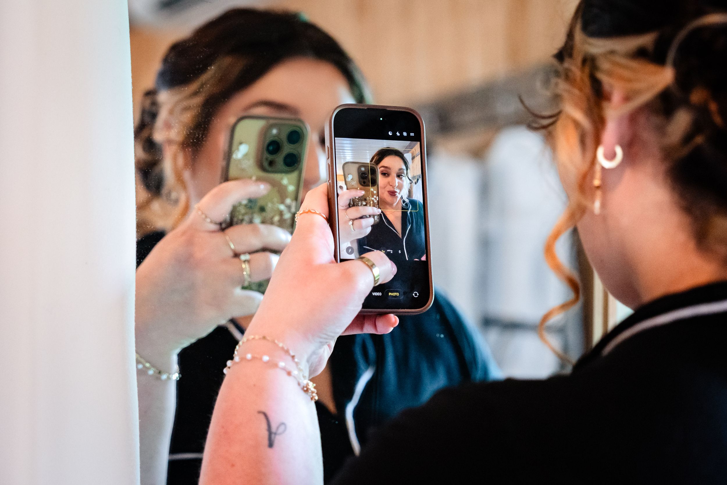 Bridesmaid photographing bride's reflection in mirror using smartphone during wedding day preparations.